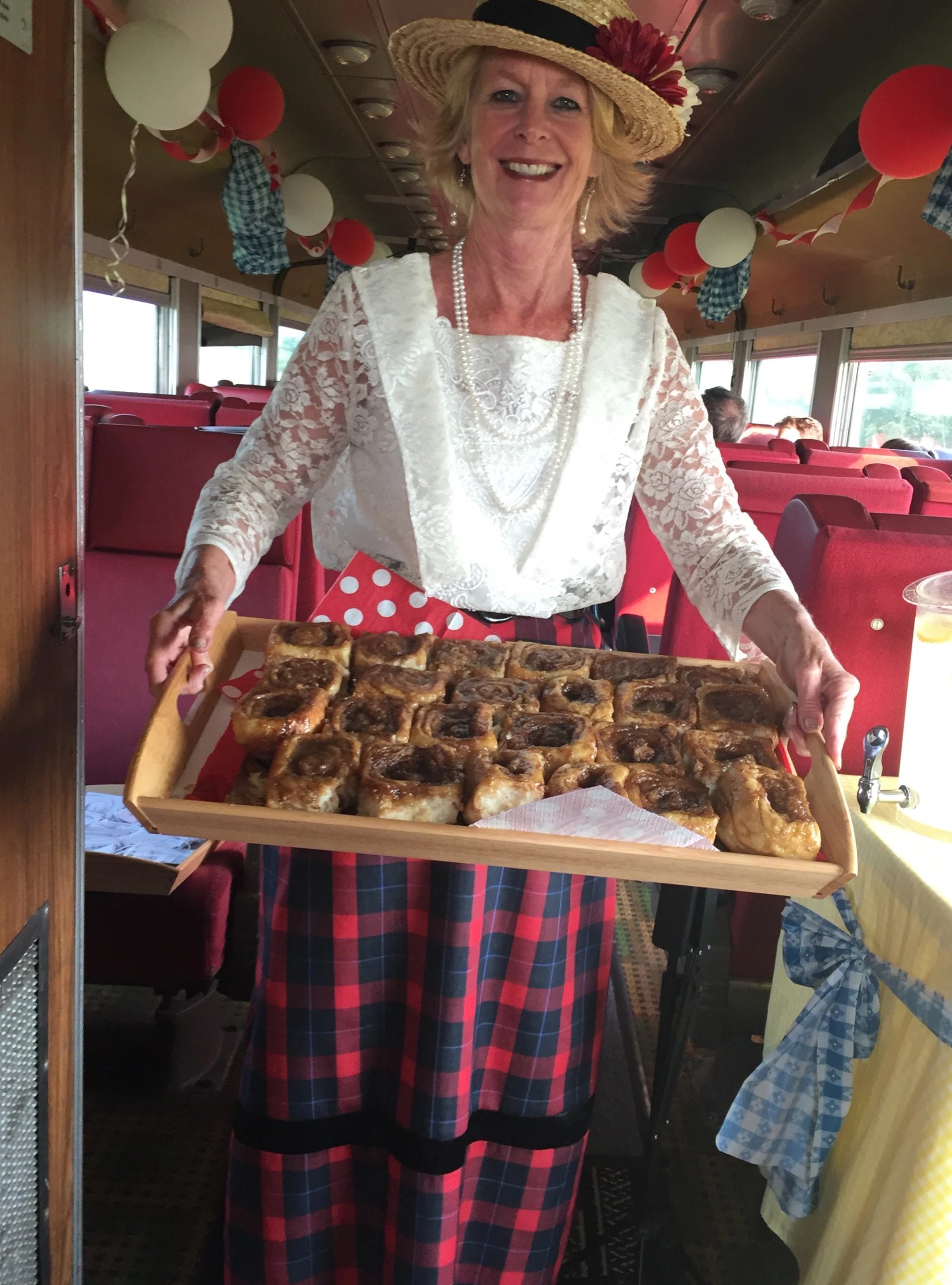 Server in lace blouse and straw hat holding tray of sticky buns on decorated train