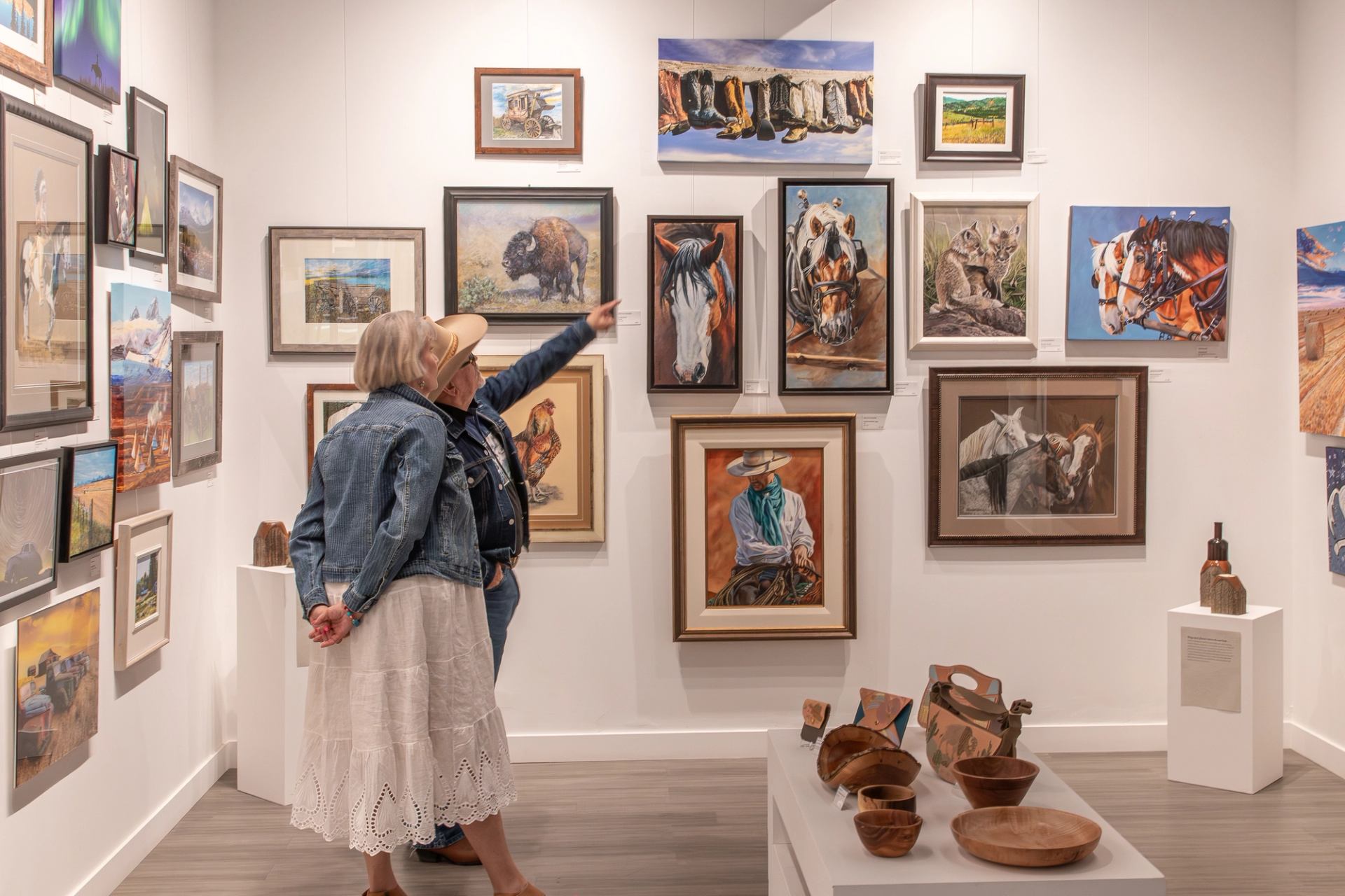 Art gallery wall with western-themed paintings as two visitors look and point.