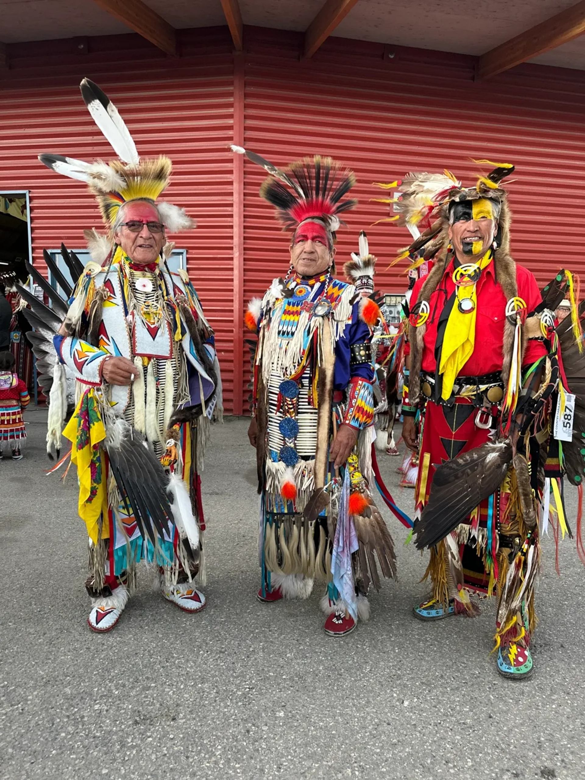 Three people in colourful traditional regalia with feathered headdresses stand outside red building.