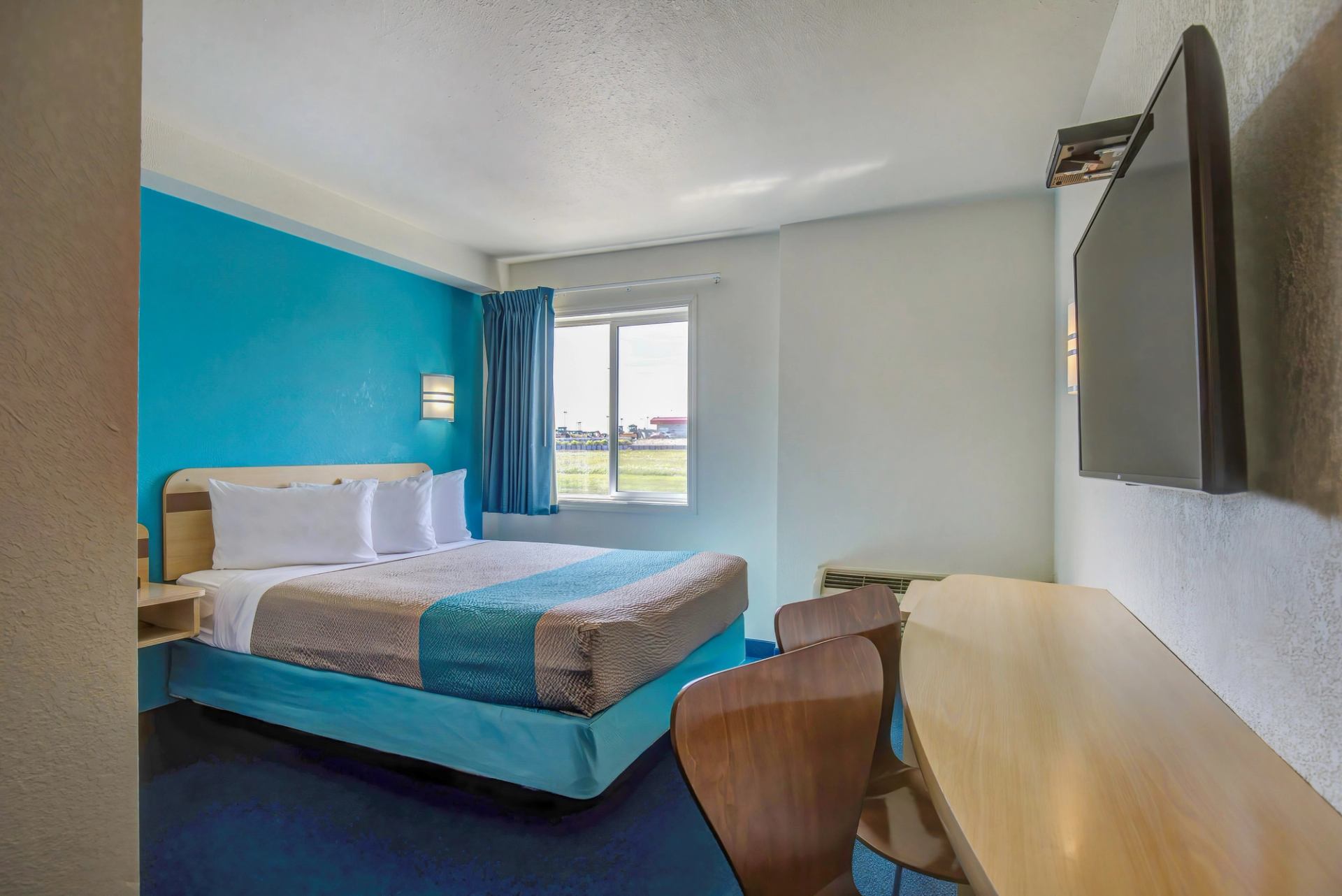 Hotel room with queen bed, blue accent wall, desk, chairs, and wall-mounted TV near window.