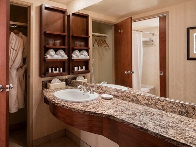 Bathroom vanity with granite countertop, sink, mirror, shelves, bathrobes and shower.
