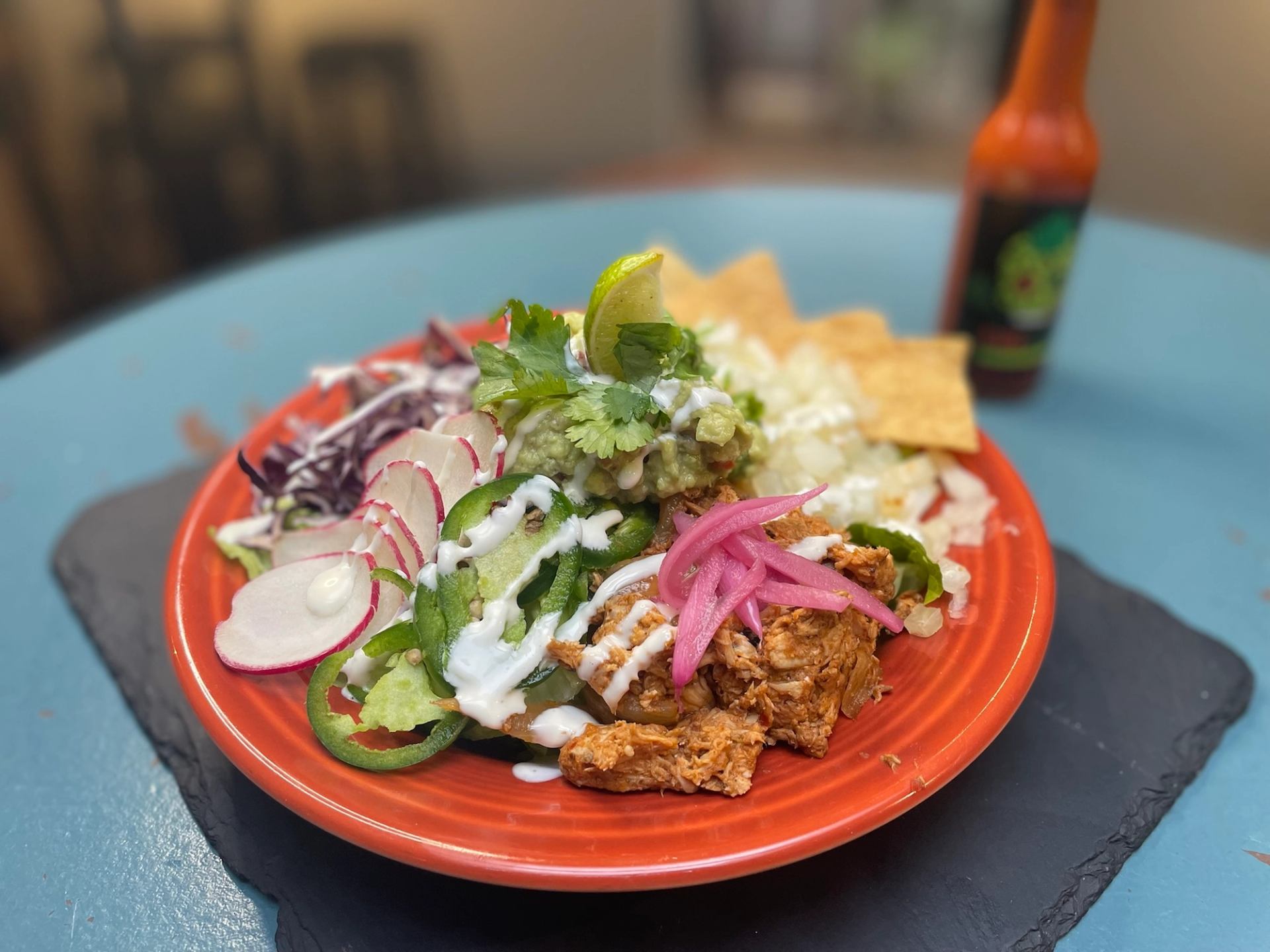 Orange plate with shredded meat, sliced vegetables, tortilla chips and hot sauce bottle.