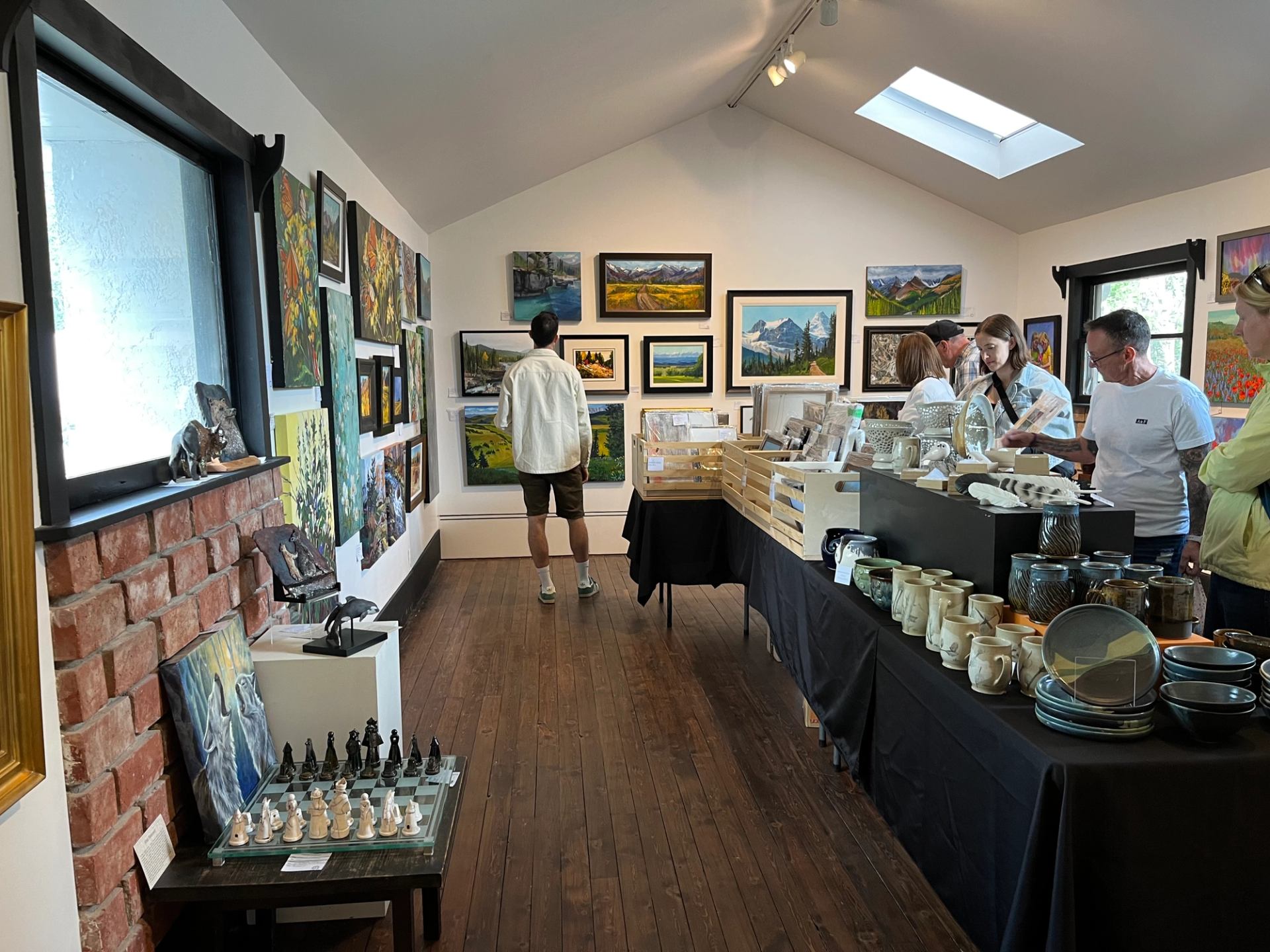 An art gallery filled with landscape paintings, a long table displaying pottery, and people browsing the artwork and crafts.