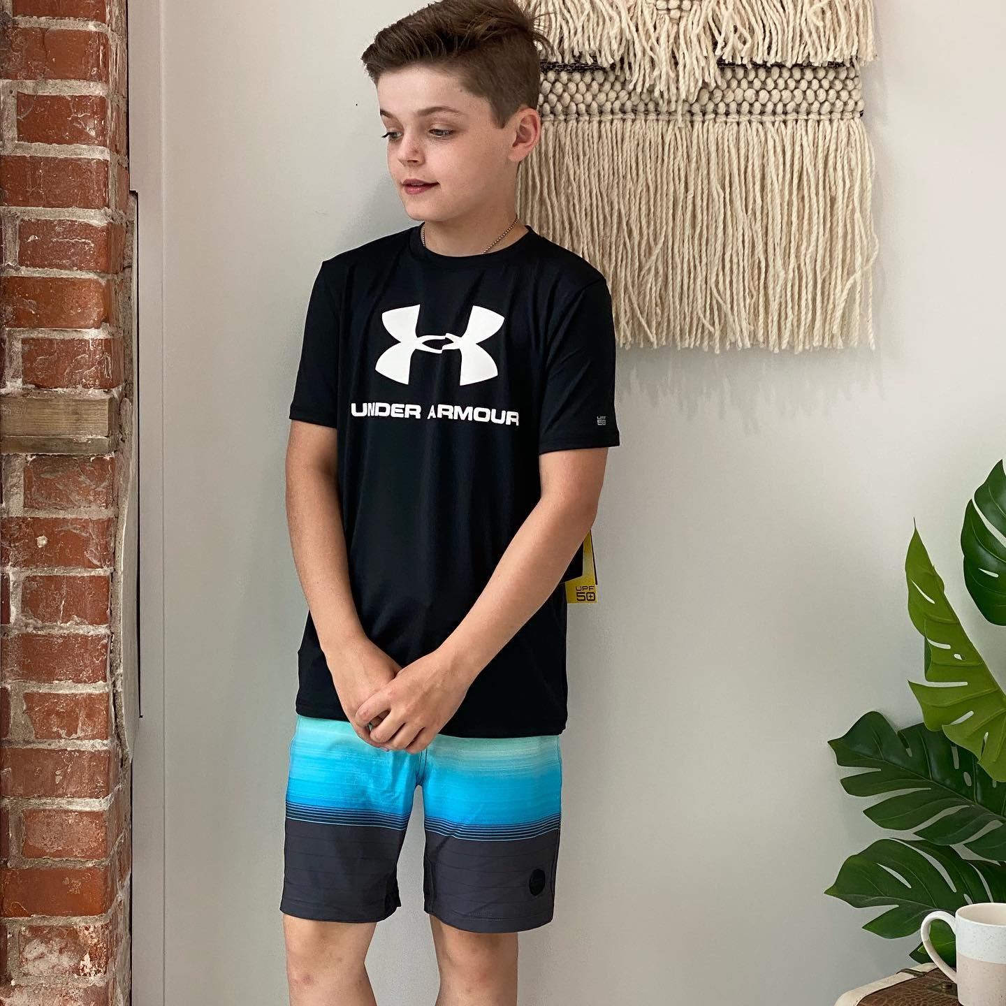 Boy wearing black Under Armour T-shirt and blue shorts, standing indoors by brick wall.