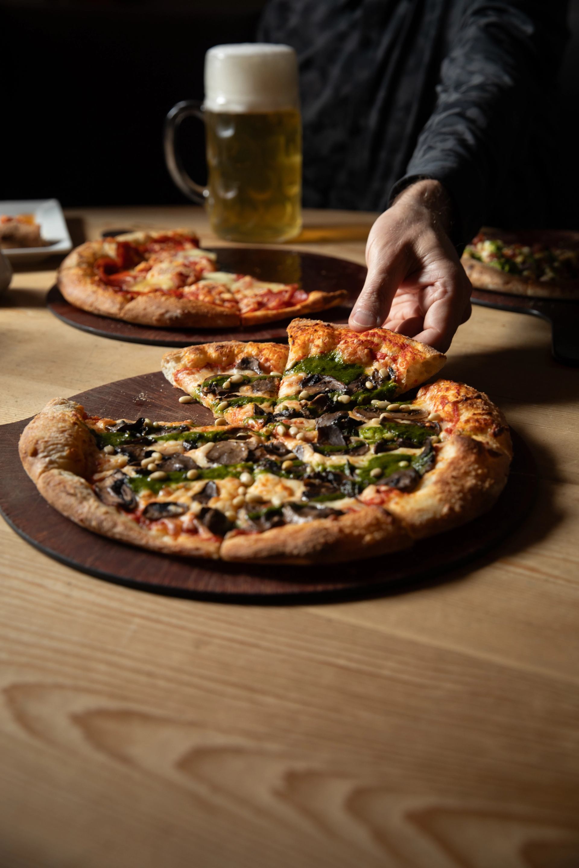 A hand grabs a slice of thin crust pizza from the table. A stein in background.