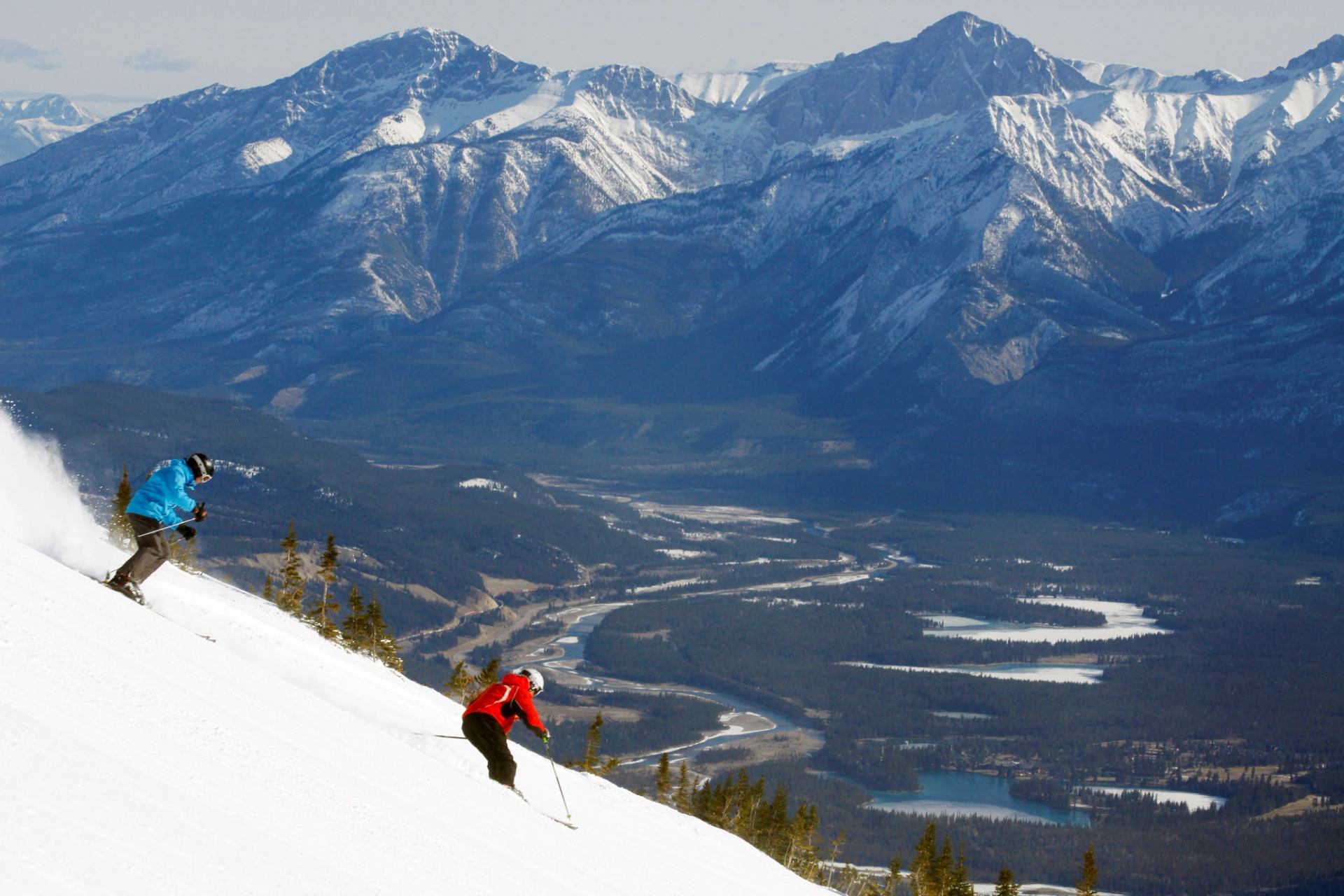 Two skiers descend a snowy slope with mountain peaks in the background.