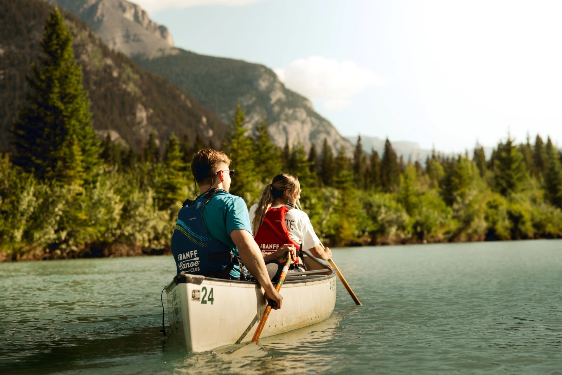 Two people paddle a canoe through a peaceful lake framed by forest and mountains in Banff.