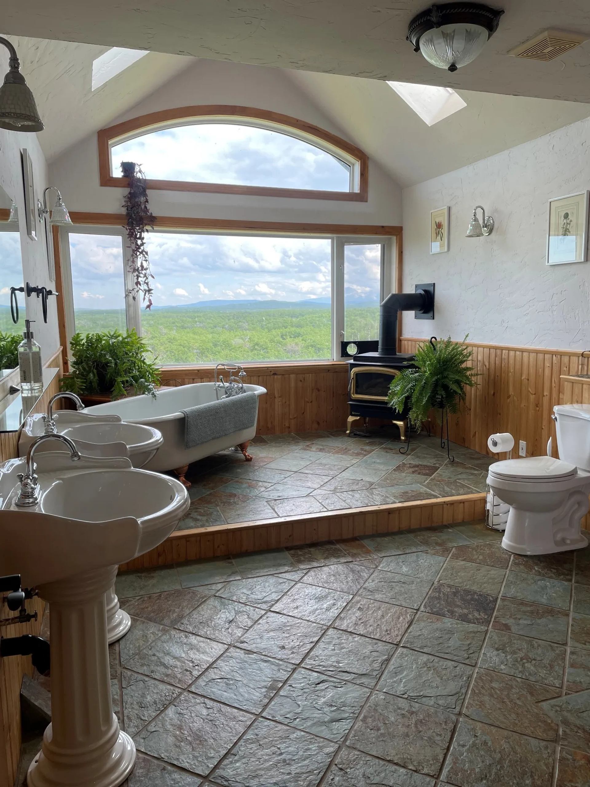 Bathroom with clawfoot tub, wood stove and large window overlooking green landscape.