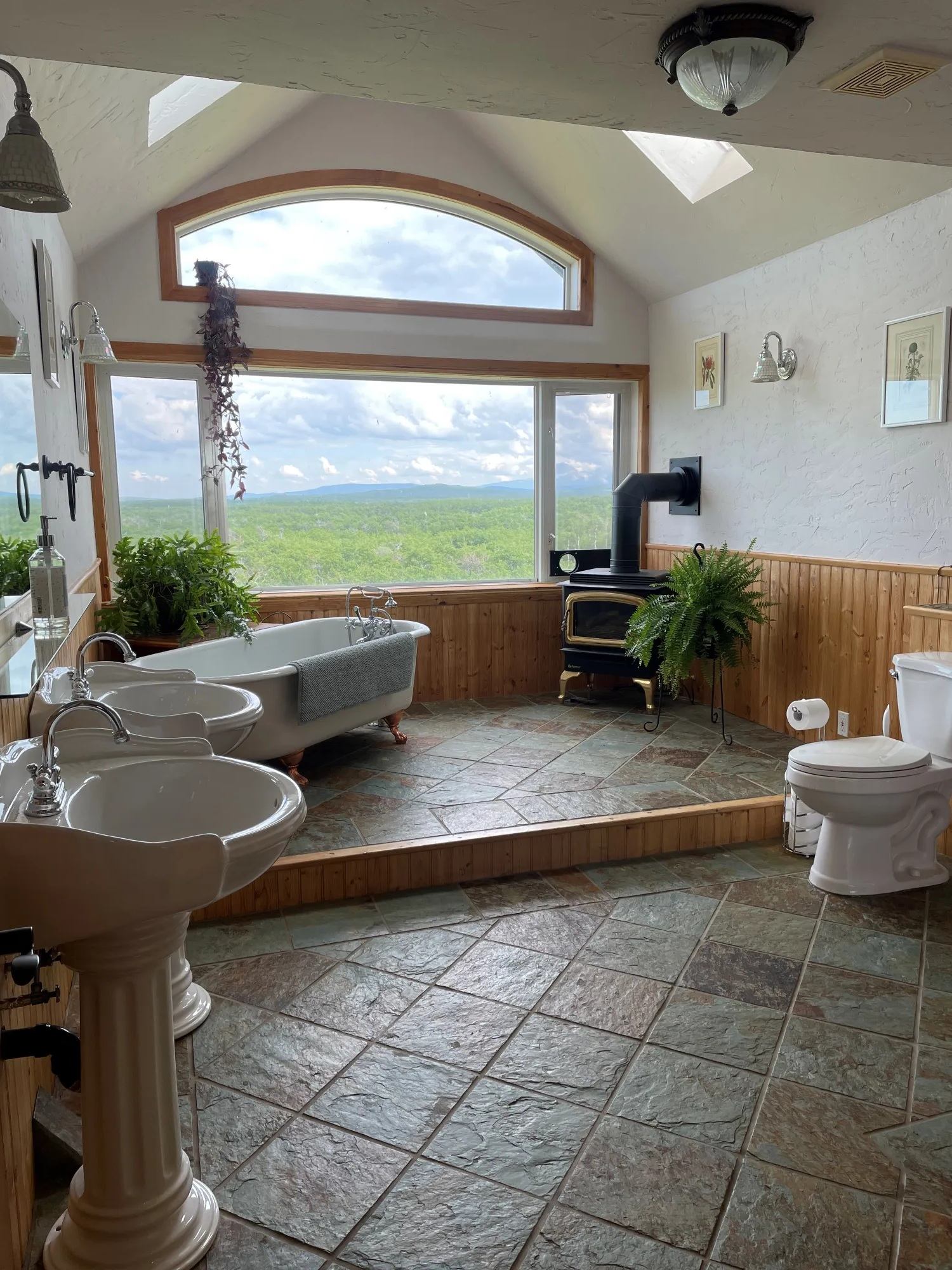 Bathroom with clawfoot tub, wood stove and large window overlooking green landscape.