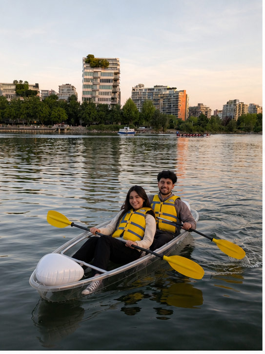 Two people paddle a clear kayak on calm water with city buildings behind.