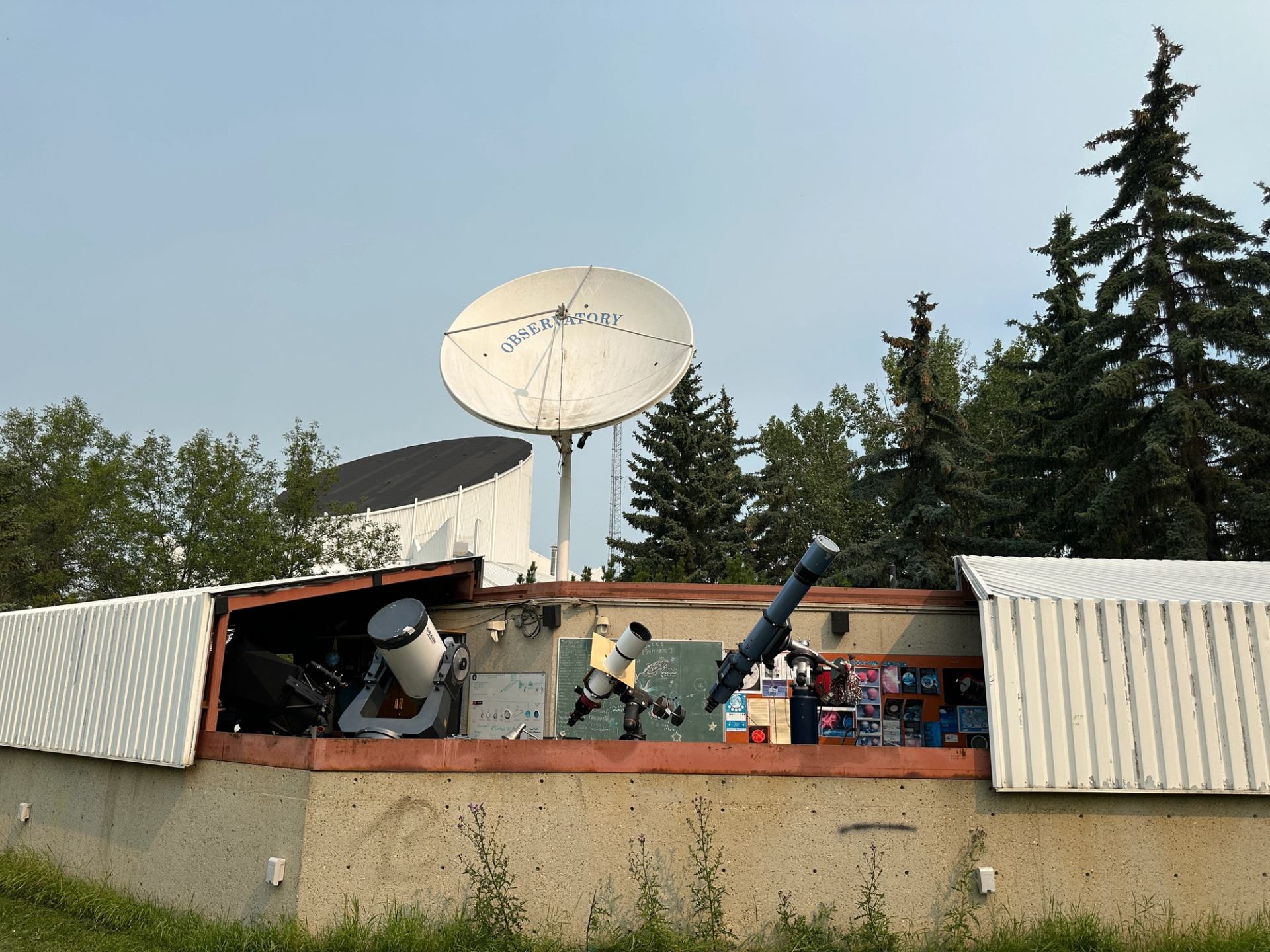 Outdoor observatory building with telescopes, satellite dish, astronomy posters and surrounding trees.