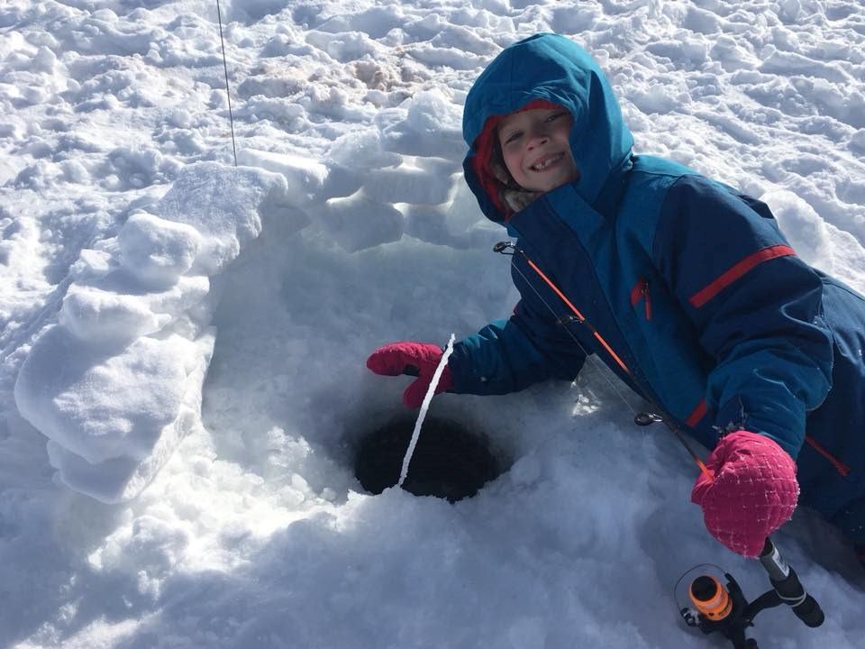 Child in blue snowsuit ice fishing beside snow shelter and hole in frozen lake.