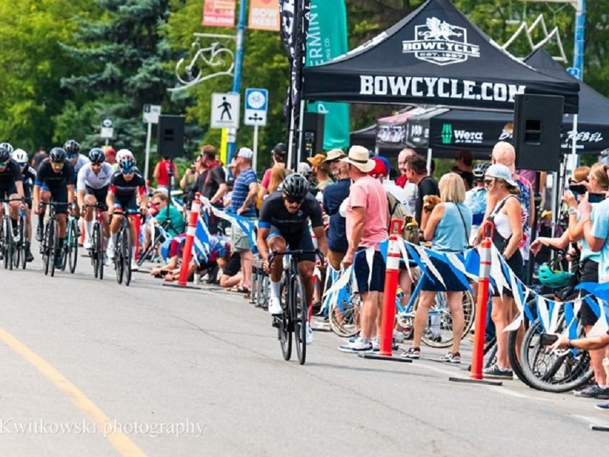 Cyclists race past spectators and Bow Cycle tent on a city street during event.