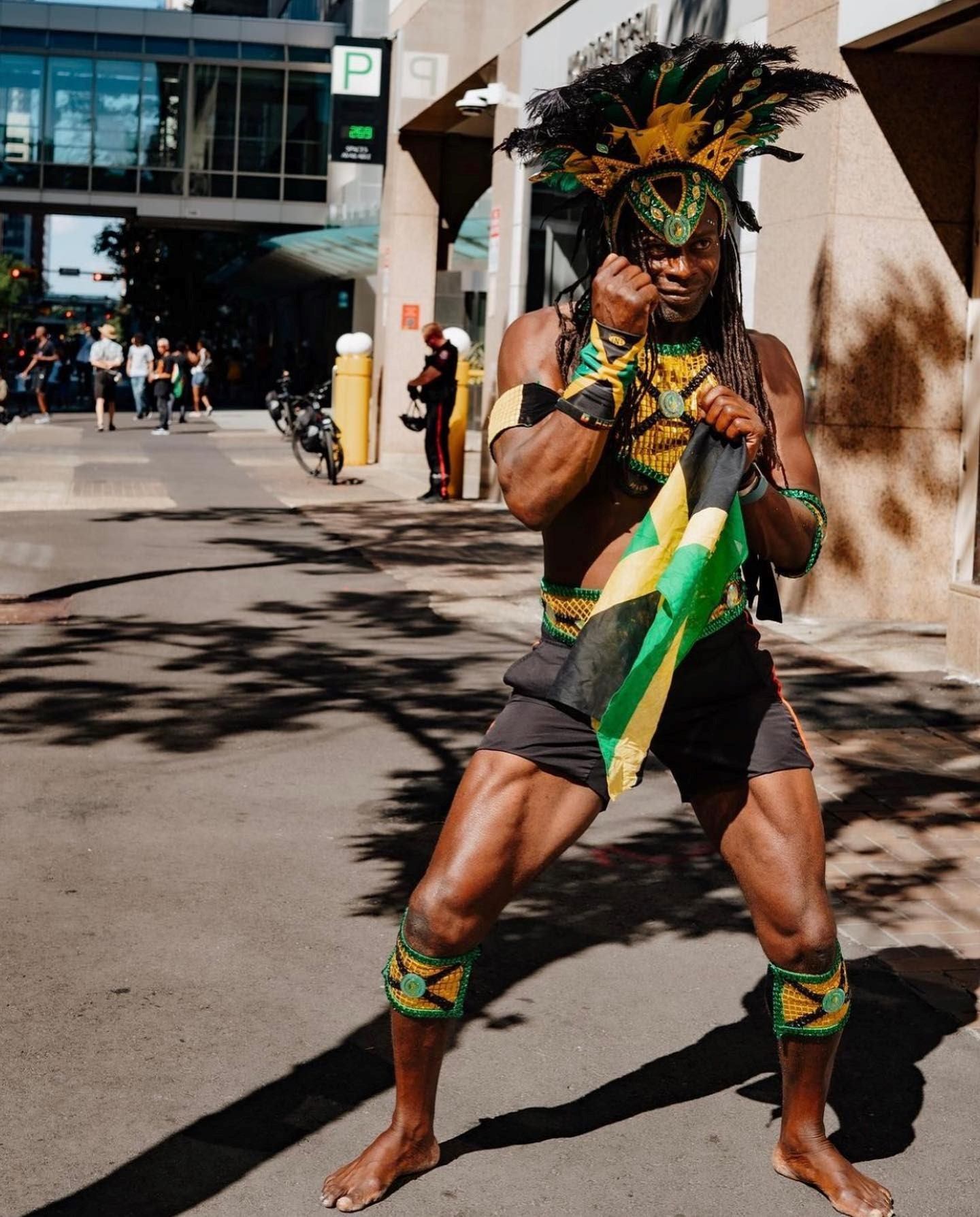 Costumed performer with feathered headdress holds Jamaican flag on city street.