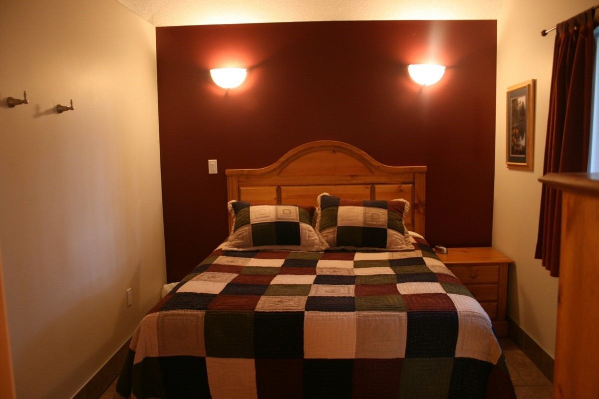 Bed with plaid quilt, wooden headboard, wall lamps, and bedside dresser in cabin bedroom.