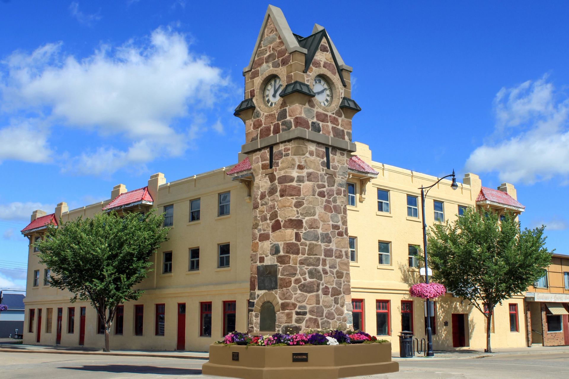 Stone clock tower with pink flowers in front of a beige building in Wainwright.