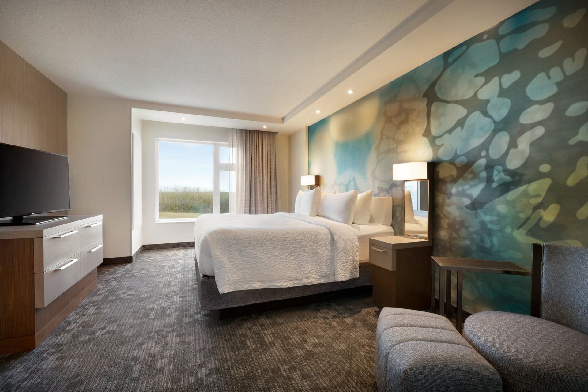 Modern hotel room with large bed, abstract mural wall, window, TV and seating.