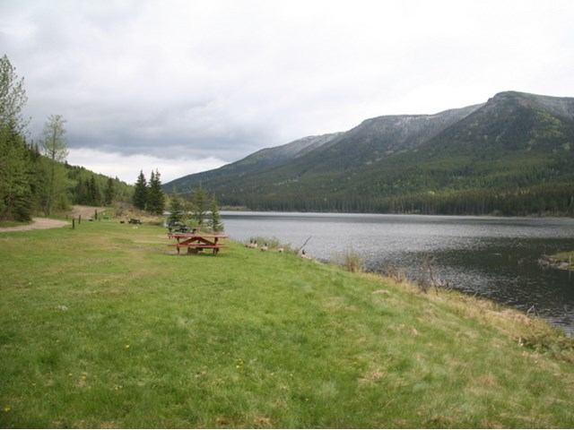 Picnic table on grassy shore overlooking a calm lake with forested mountains in the background.