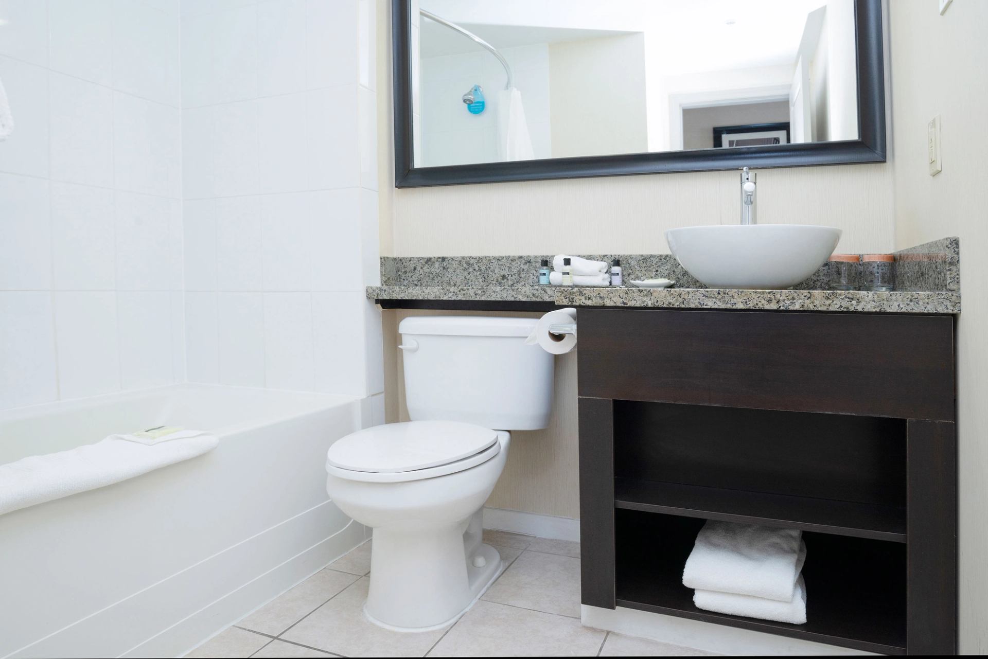 Bathroom with bathtub, toilet, granite vanity, vessel sink and folded towels on shelf.
