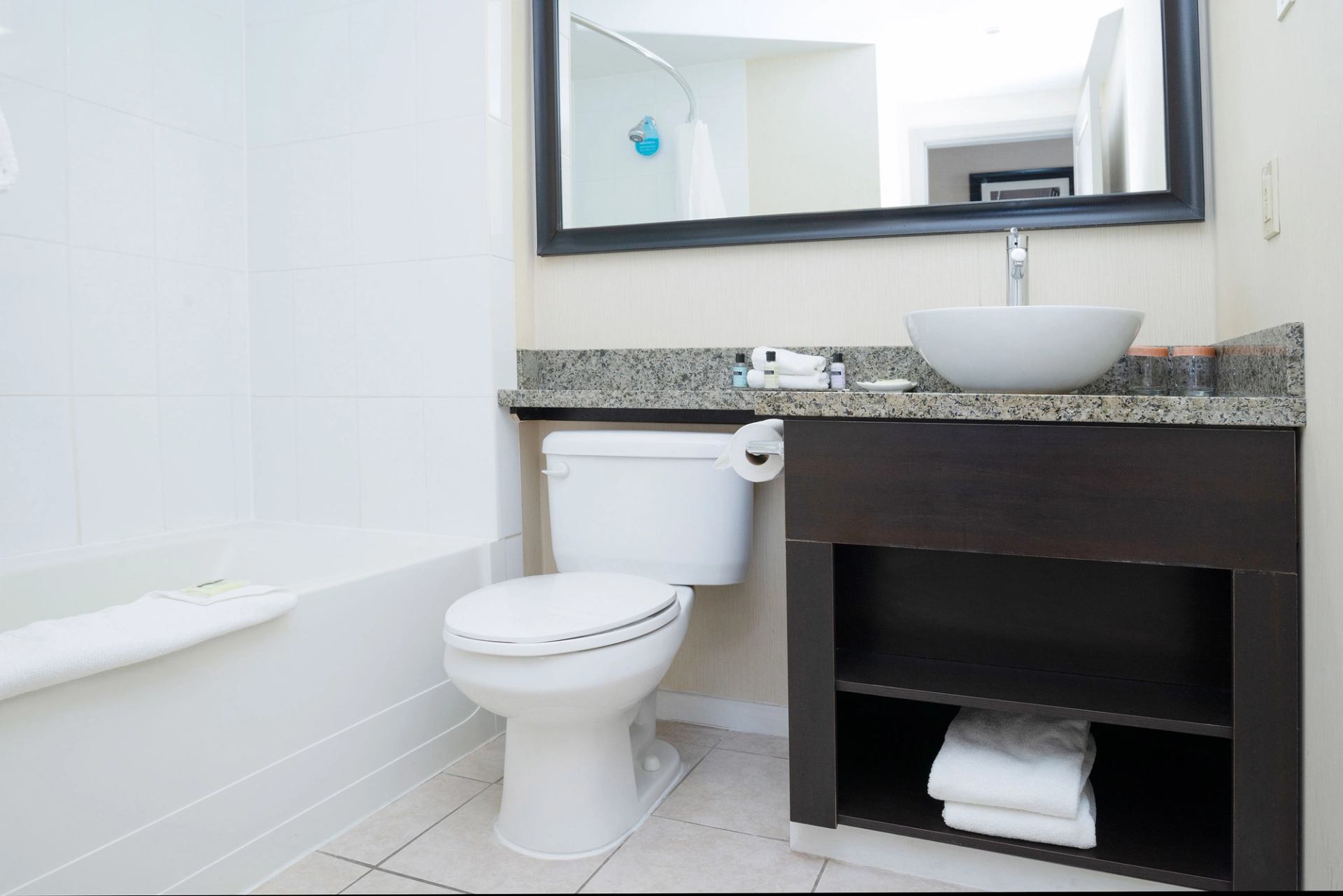 Bathroom with bathtub, toilet, granite vanity, vessel sink and folded towels on shelf.