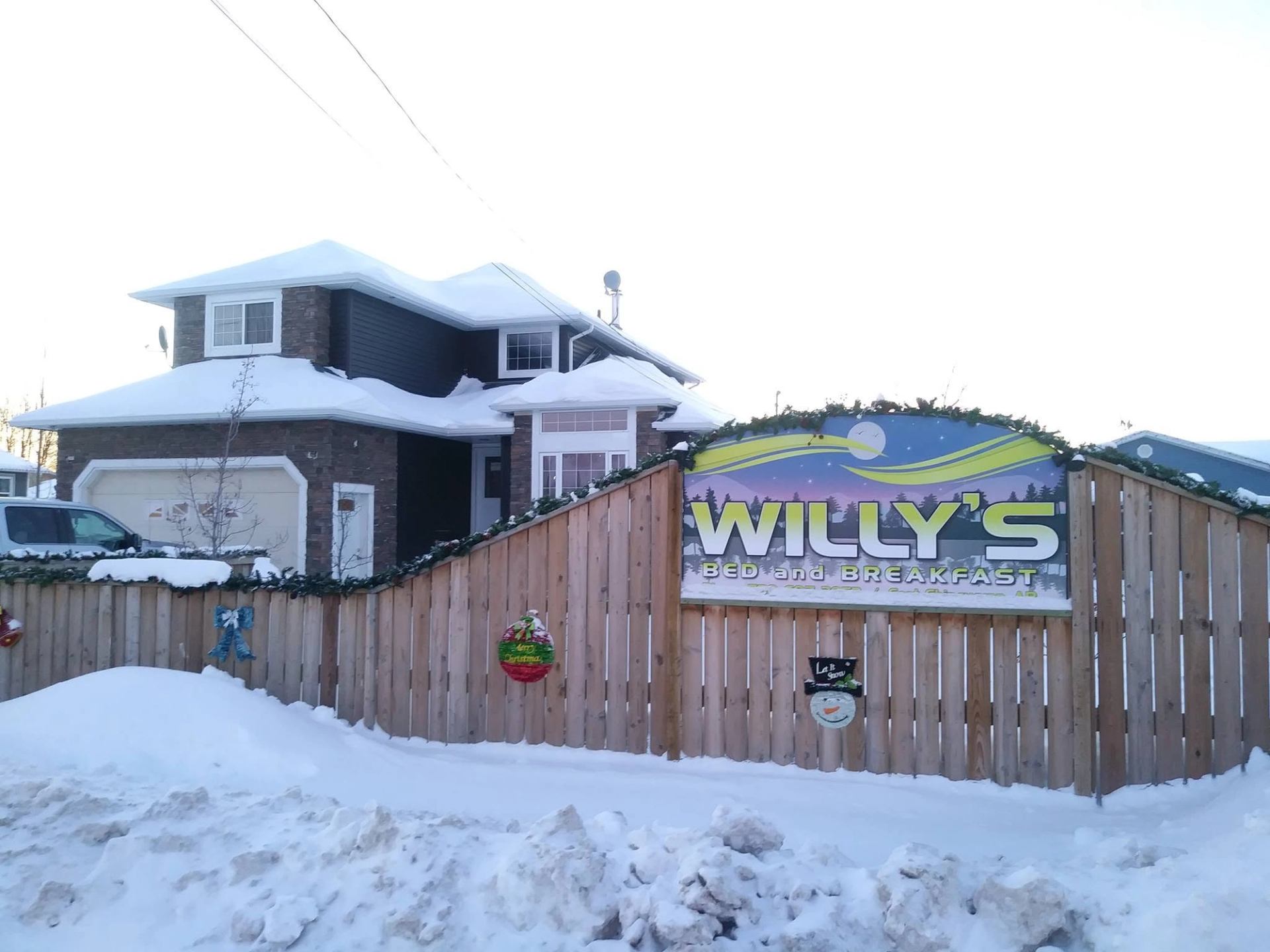 Snow-covered house behind wooden fence with Willy’s Bed and Breakfast sign and decorations.