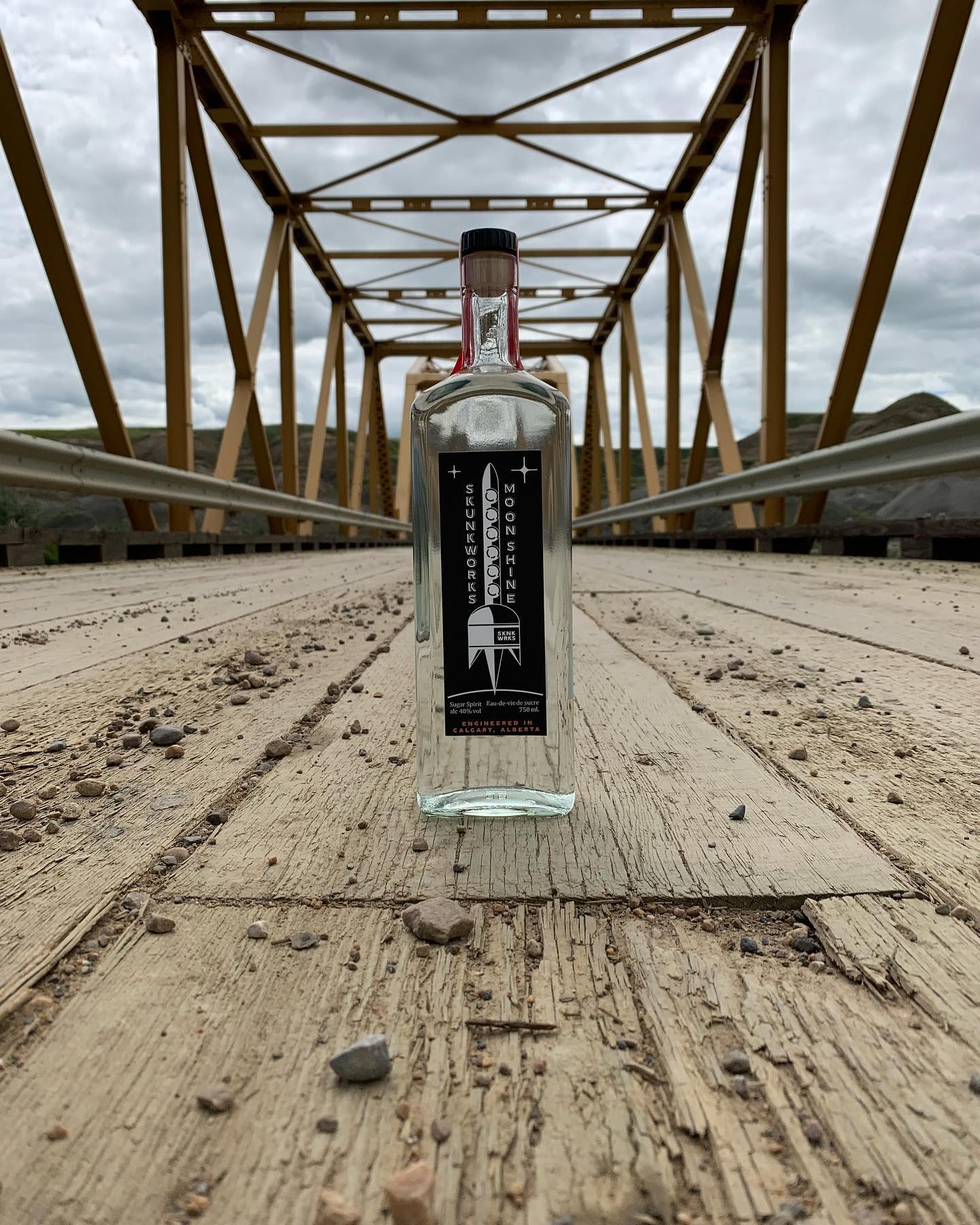 Clear Skunkworks Moonshine bottle standing on weathered wooden bridge with metal trusses.