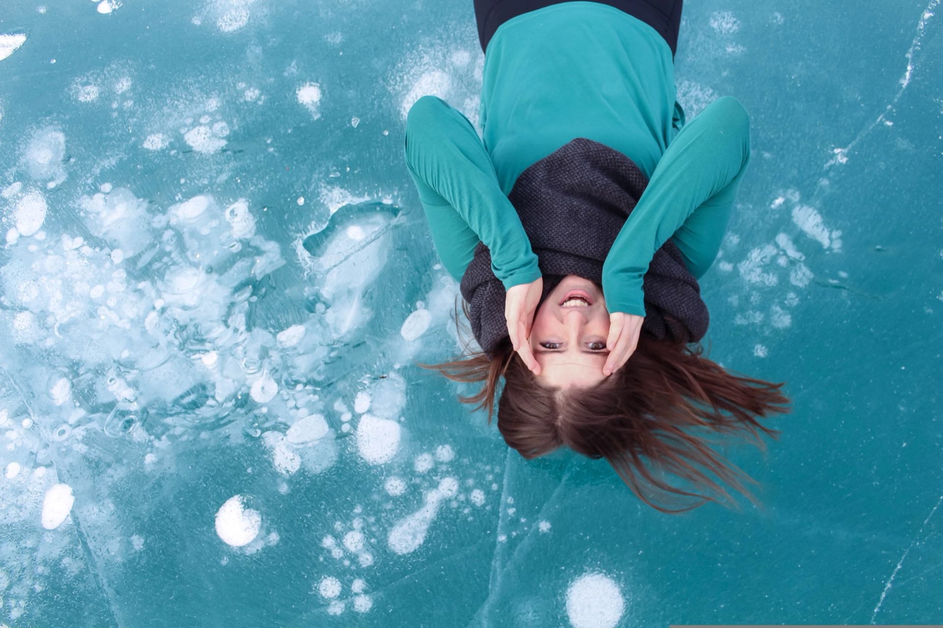 Person in teal clothing lies on blue ice with hands framing their face.