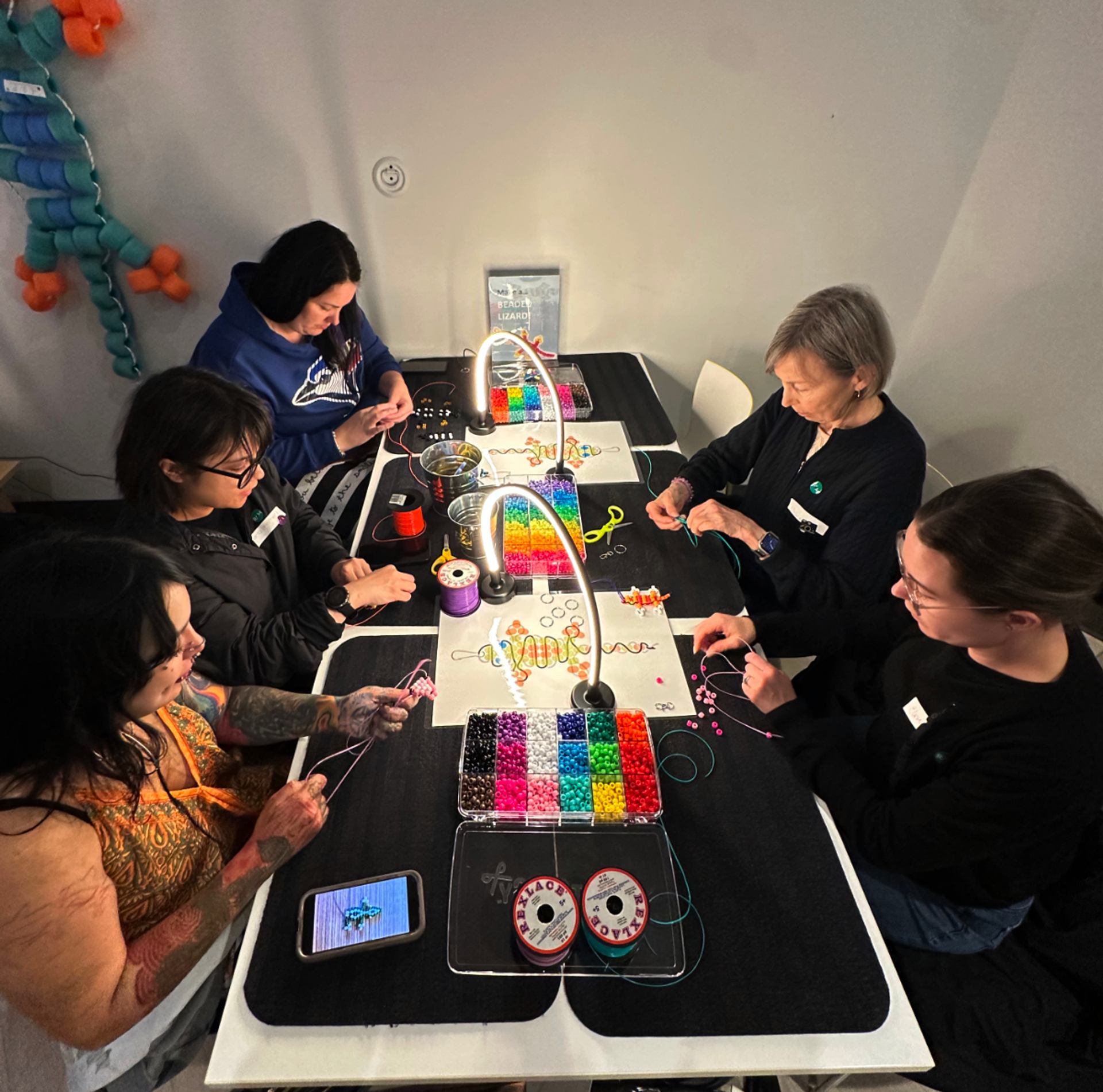 People make jewellery around a table covered with colourful beads, cords and tools.