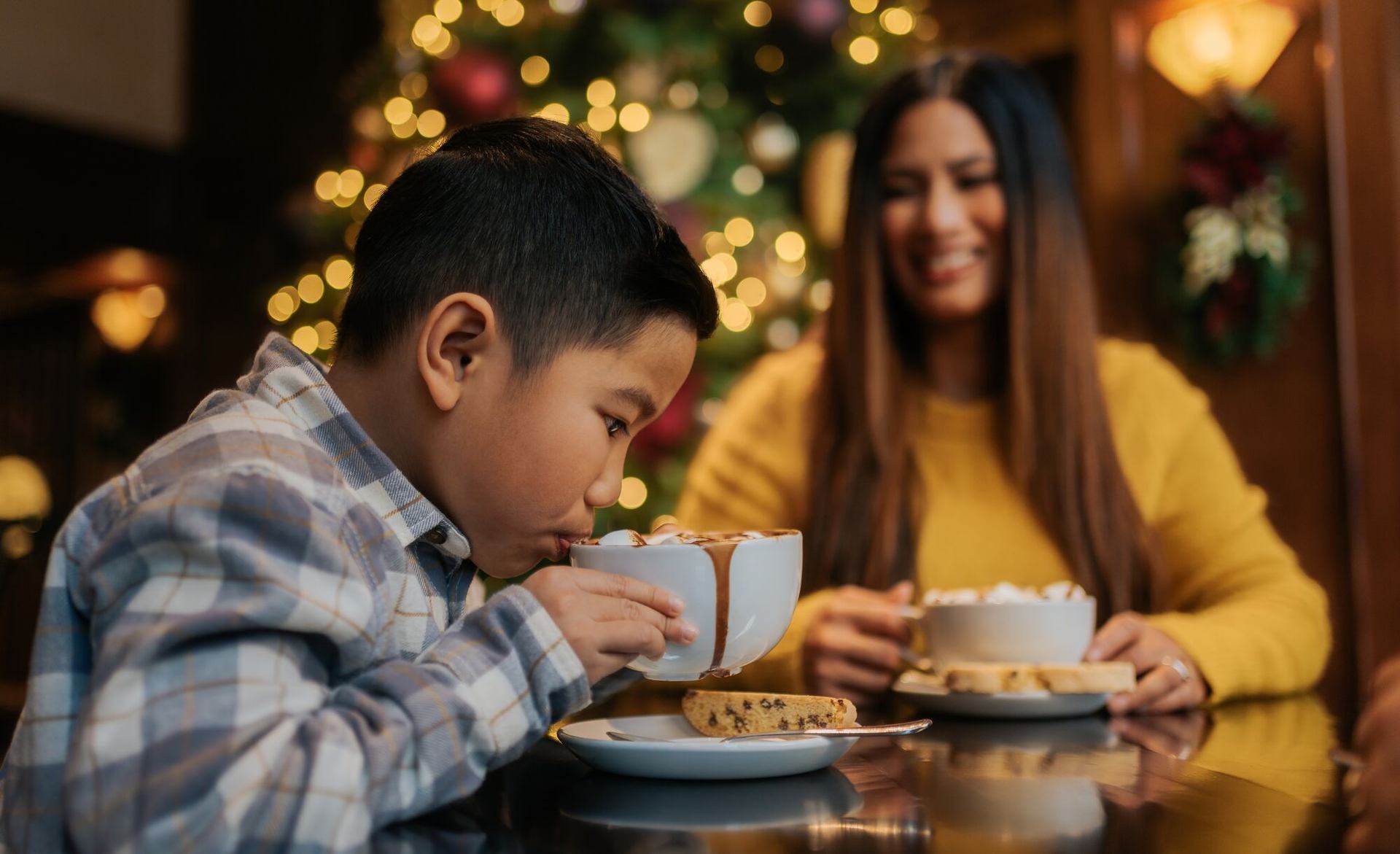 Child and adult enjoying hot chocolate with cookies near a festive Christmas tree.