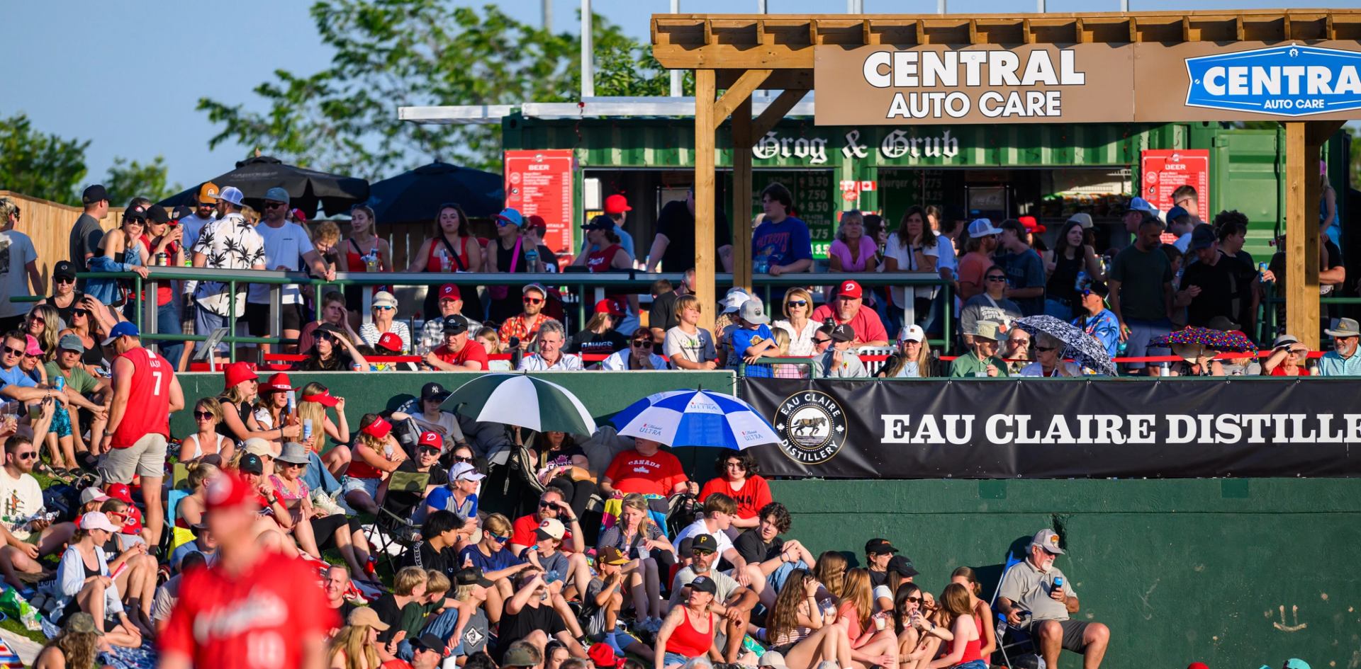 Fans gathered in sunny stands at a baseball game with banners and umbrellas.