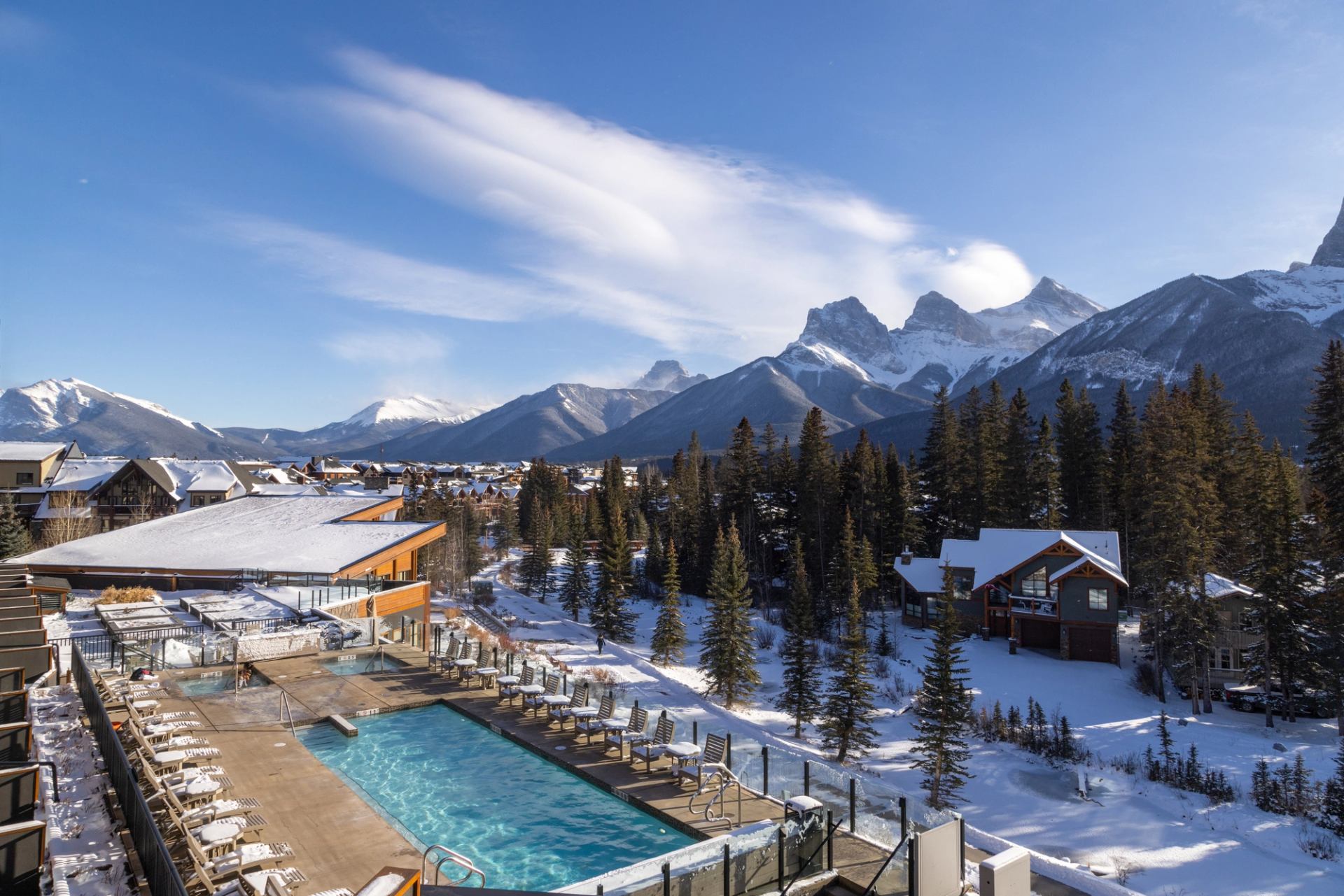 Outdoor pool overlooks snow-covered evergreens, houses and rugged mountain peaks.
