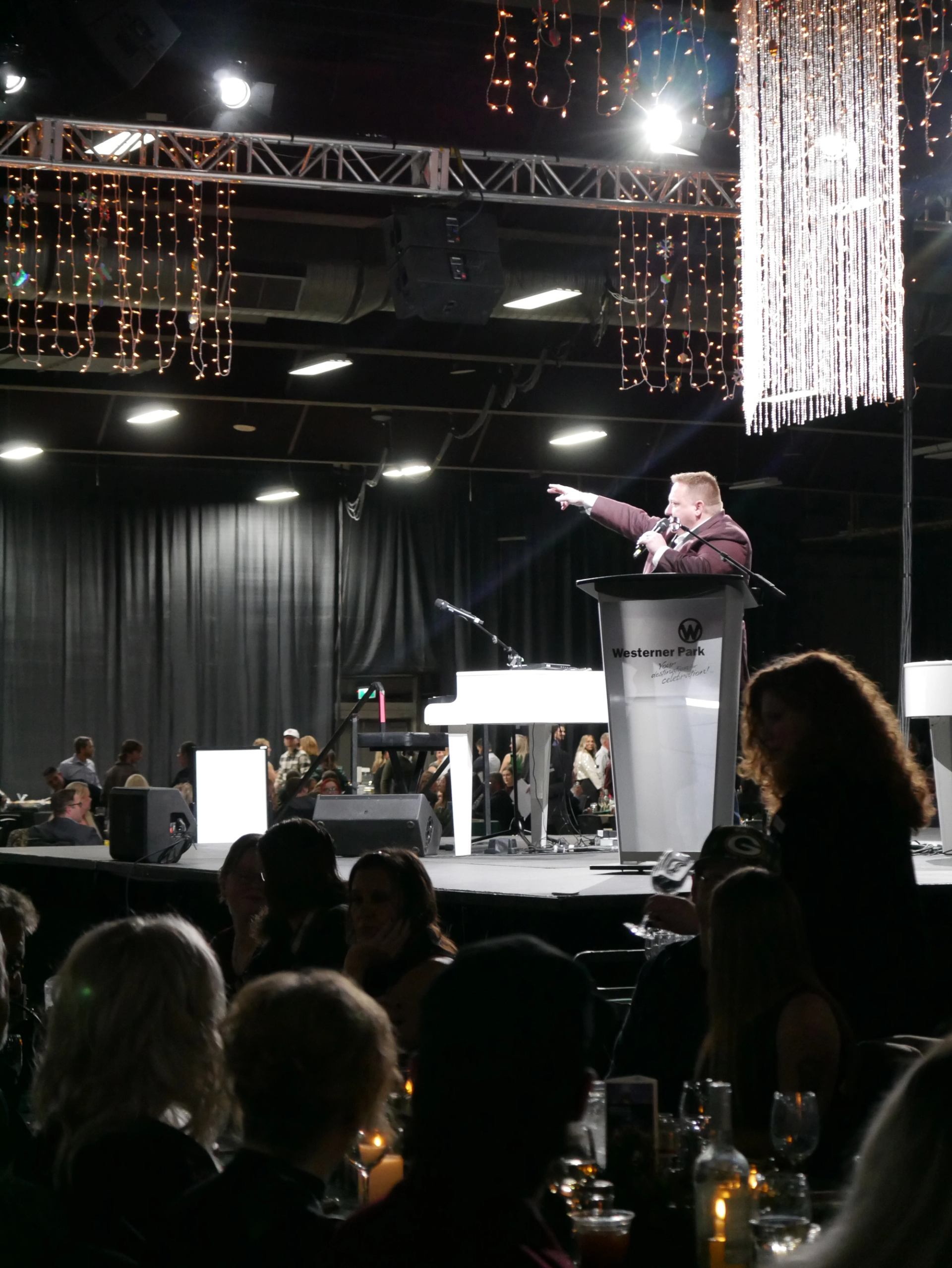 Speaker addresses a seated audience from a Westerner Park podium beneath hanging lights.