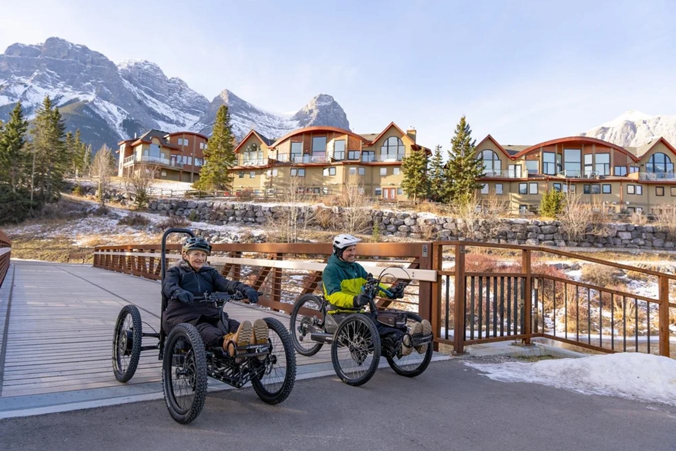 Two people ride adaptive quad bikes on a bridge below snow-capped mountains and townhouses.