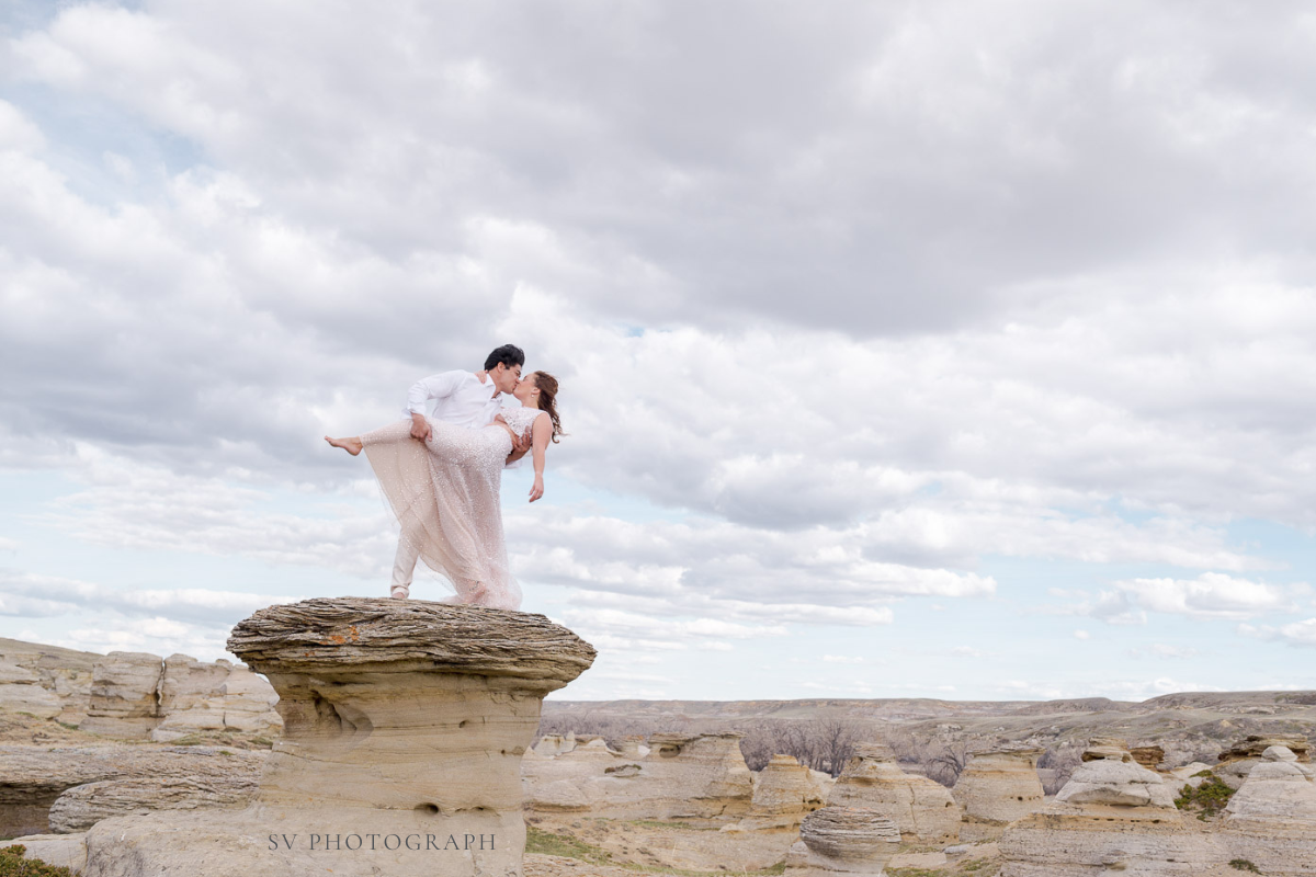 A couple stands atop a layered rock formation, one lifting the other beneath a wide, cloud-filled sky.