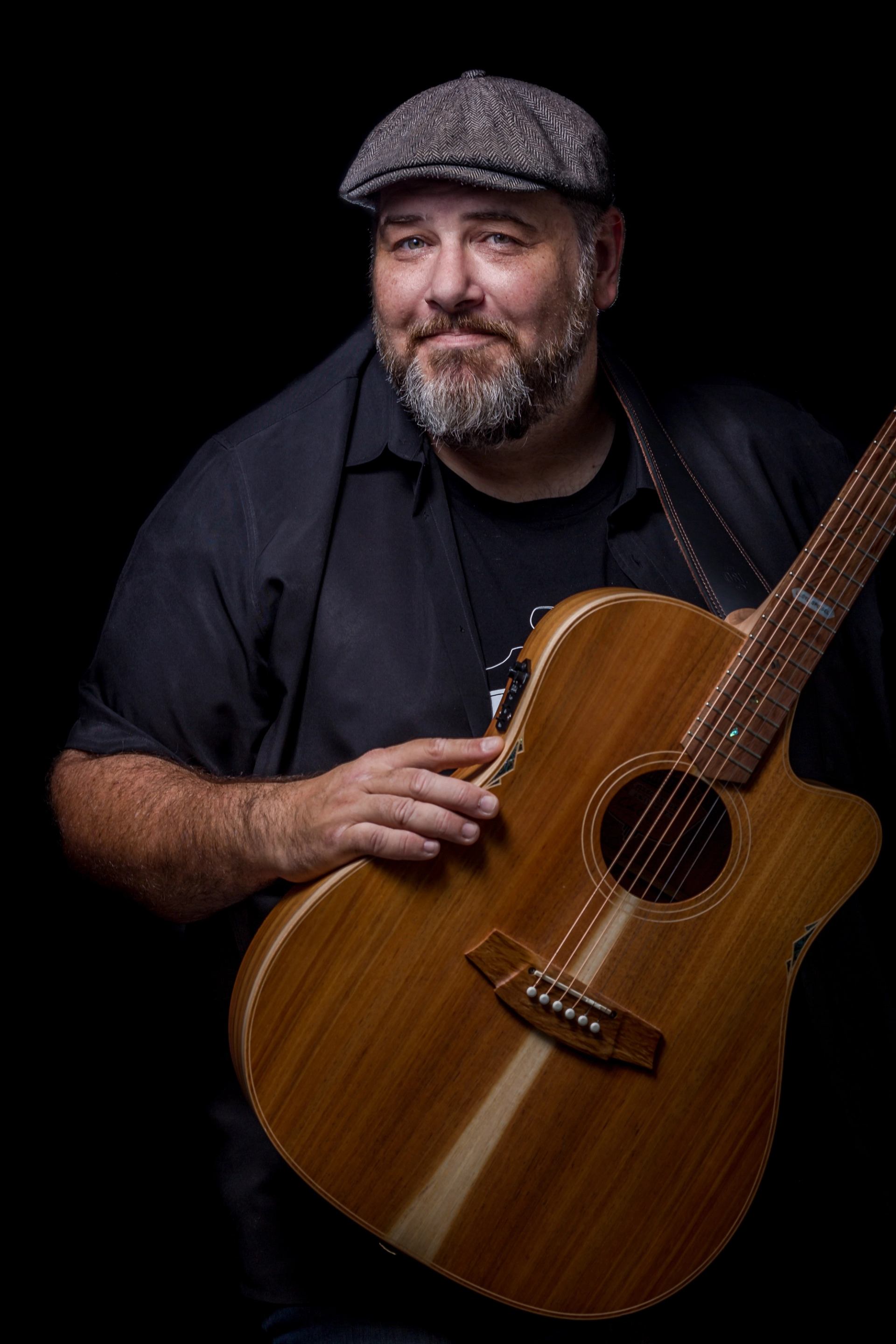 Person in flat cap holds an acoustic guitar against a dark studio background.