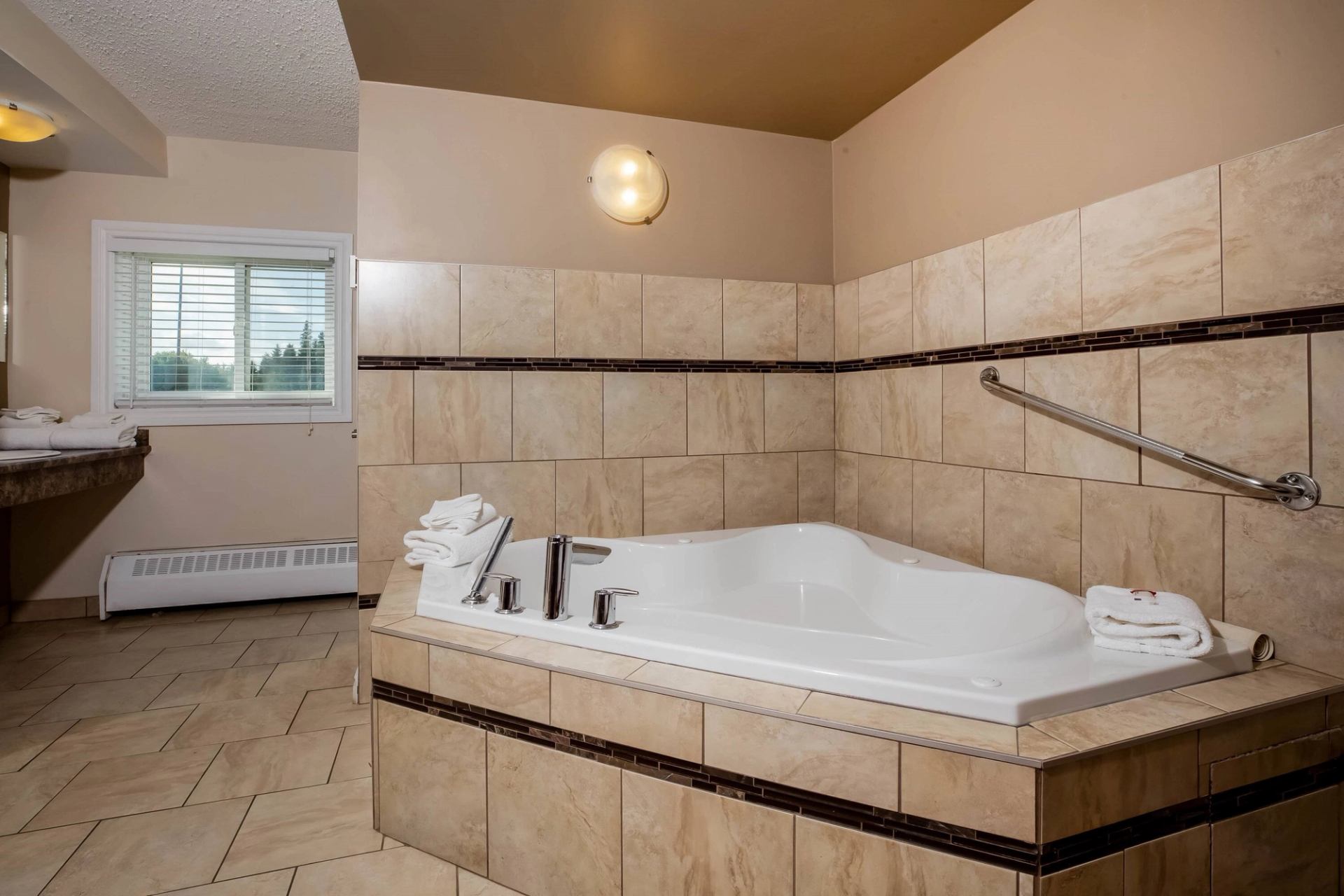 A jacuzzi bathtub surrounded by beige tiles, with a window, towel racks, and modern lighting in the room.