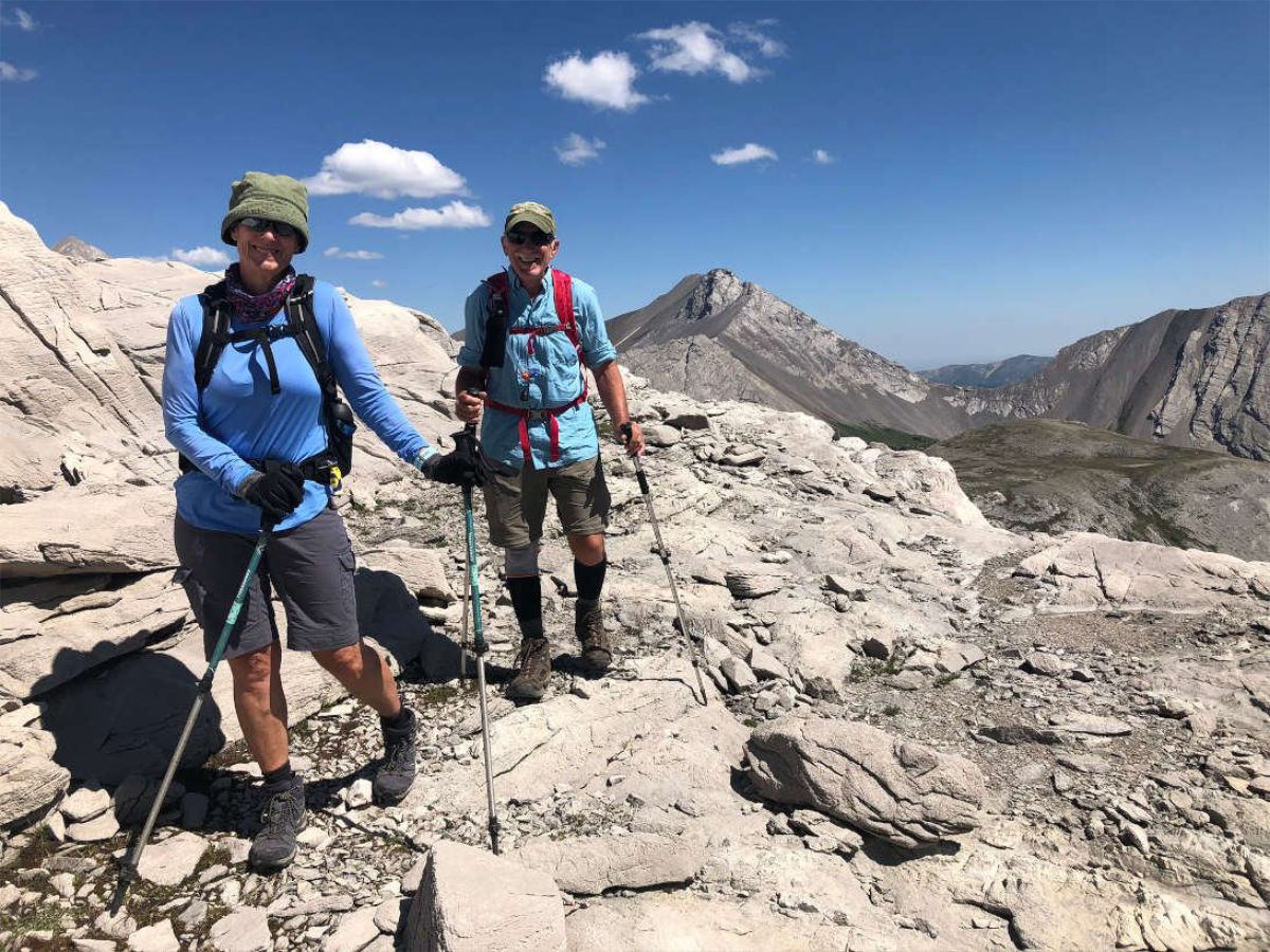 Hikers with trekking poles walk across rocky mountain ridge under clear blue sky.