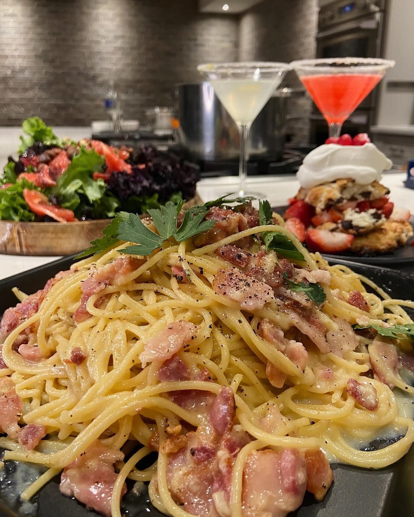 Creamy pasta with bacon, fresh salad, and cocktails on a modern kitchen counter.