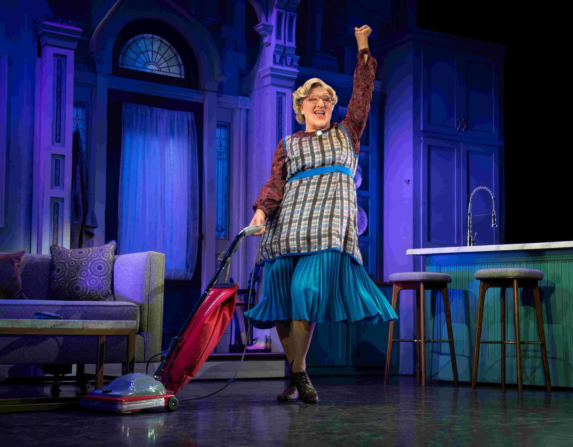 Stage performer in apron vacuums beside living room sofa and kitchen island under blue lighting..