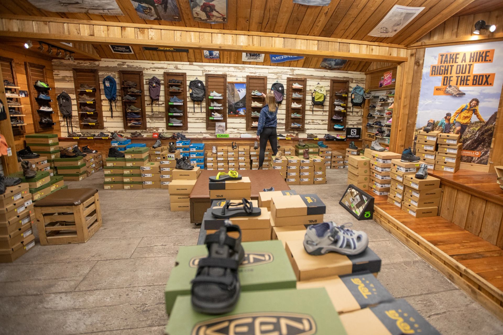 Outdoor shoe store interior with stacked boxes, wall displays and one shopper browsing.