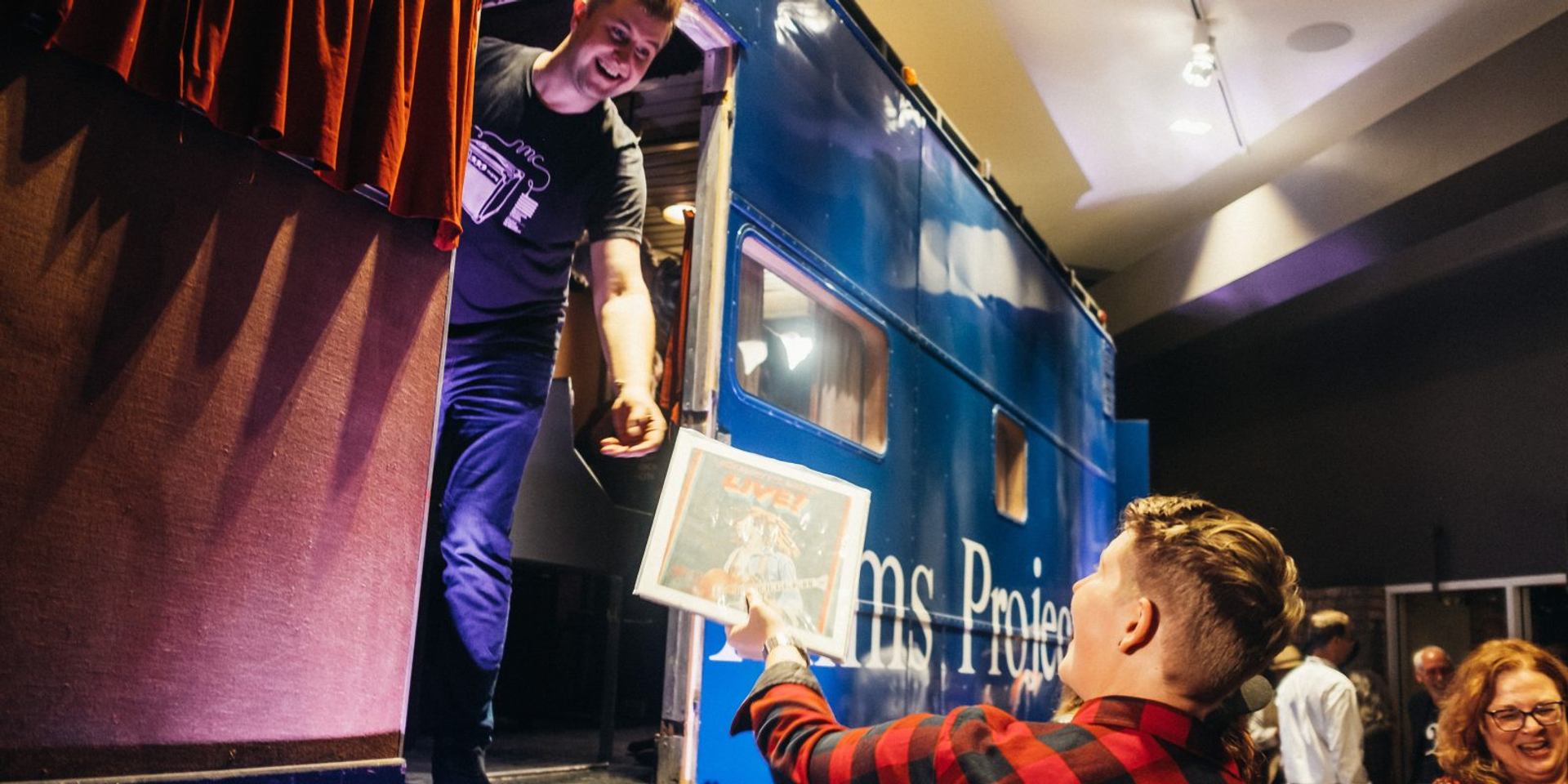 Man on blue truck stage reaches for framed poster from audience member.