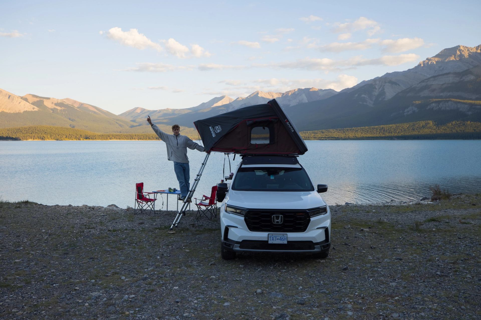 SUV with rooftop tent by a lake, mountains in background, person raising arm near camping setup.