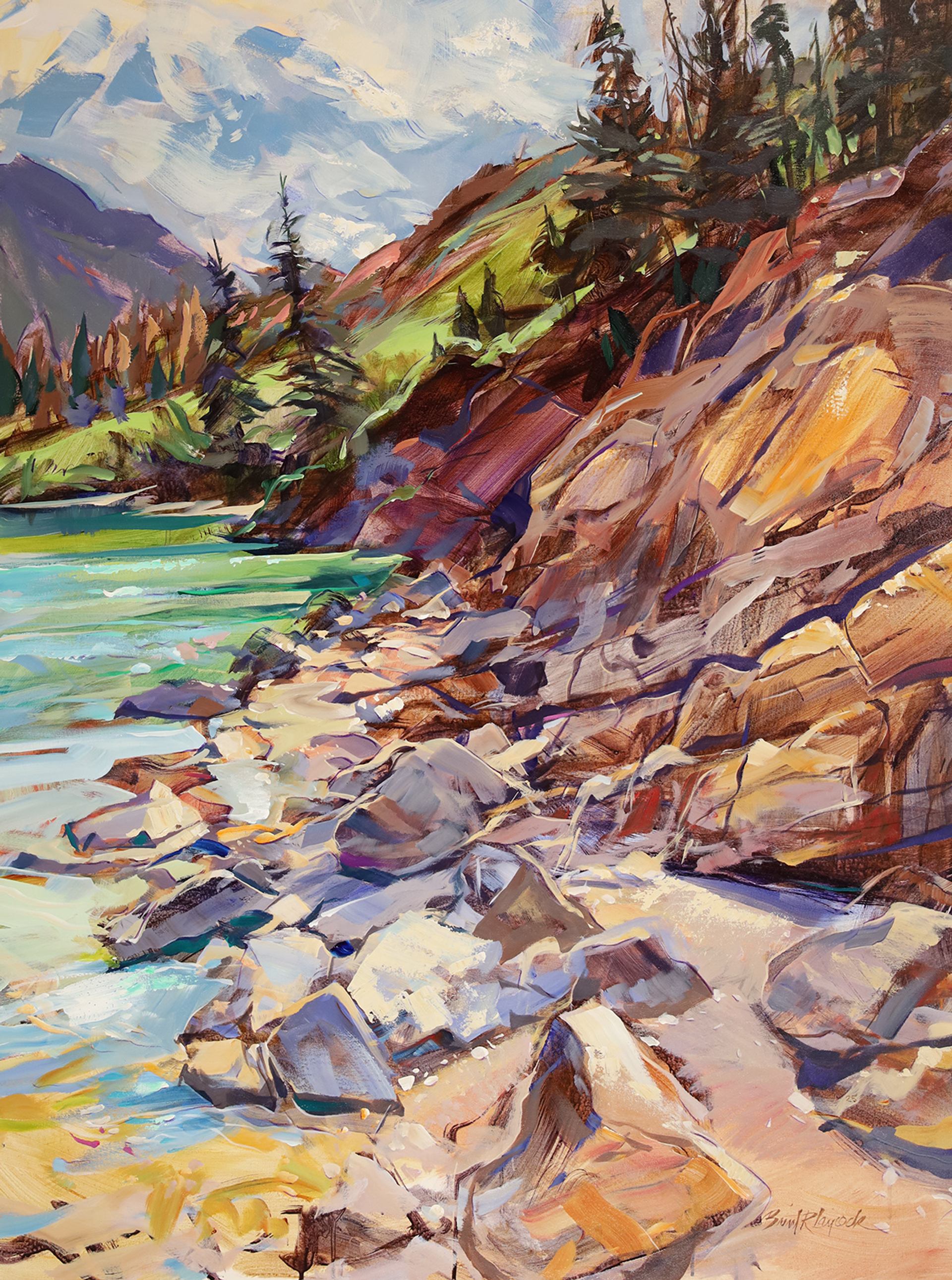 Colorful rocky shoreline with evergreens, turquoise water, and distant mountains.