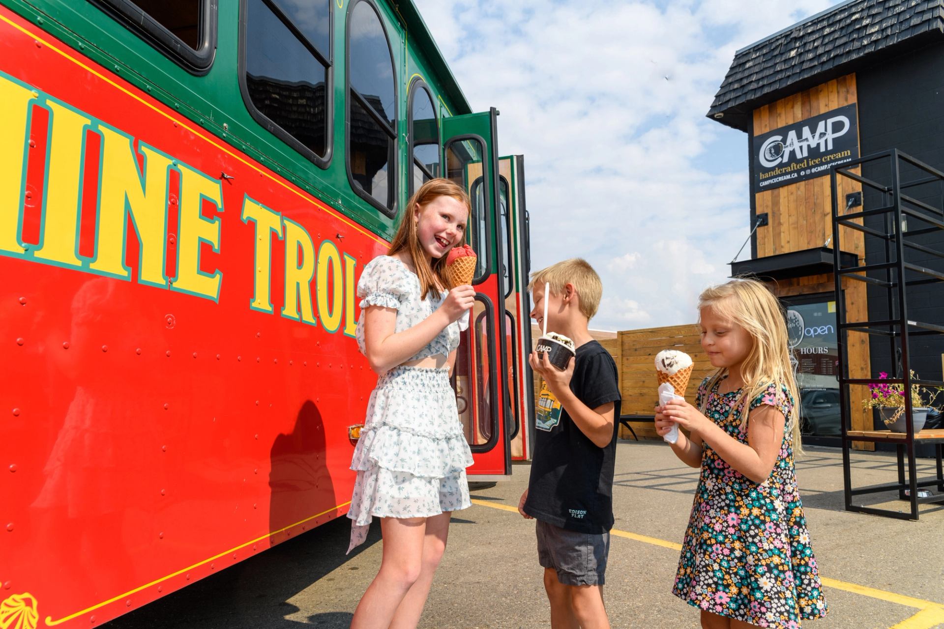 Kids eating ice cream cones beside a colorful trolley and an ice cream shop.