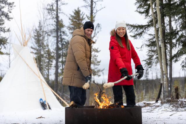 A couple cooking bannock by fire, tipi in the background, at the Aurora Borealis Indigenous Village.