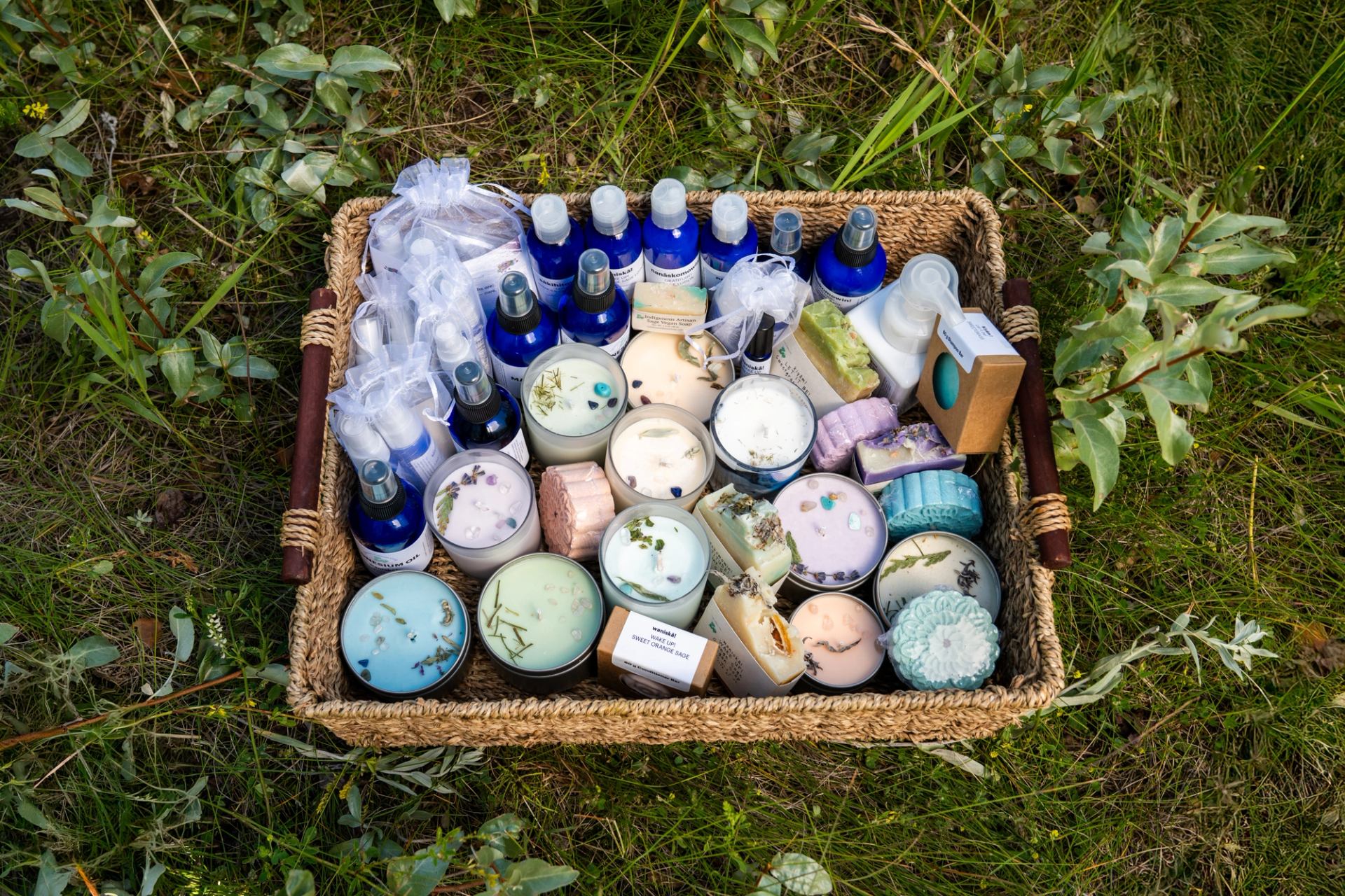 A woven basket holds candles, sprays, soaps, and bottles arranged on grass.