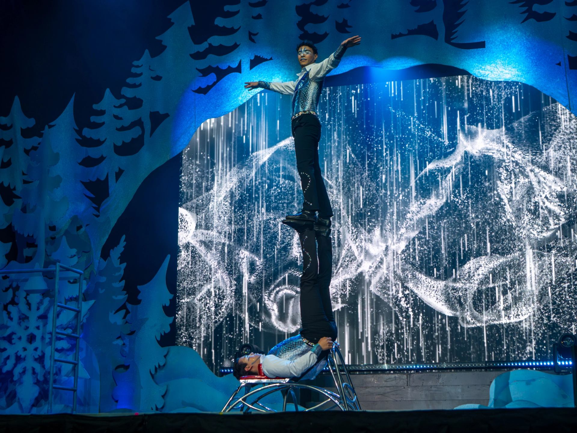 Acrobats balance vertically on a chair against an illuminated mountain backdrop.
