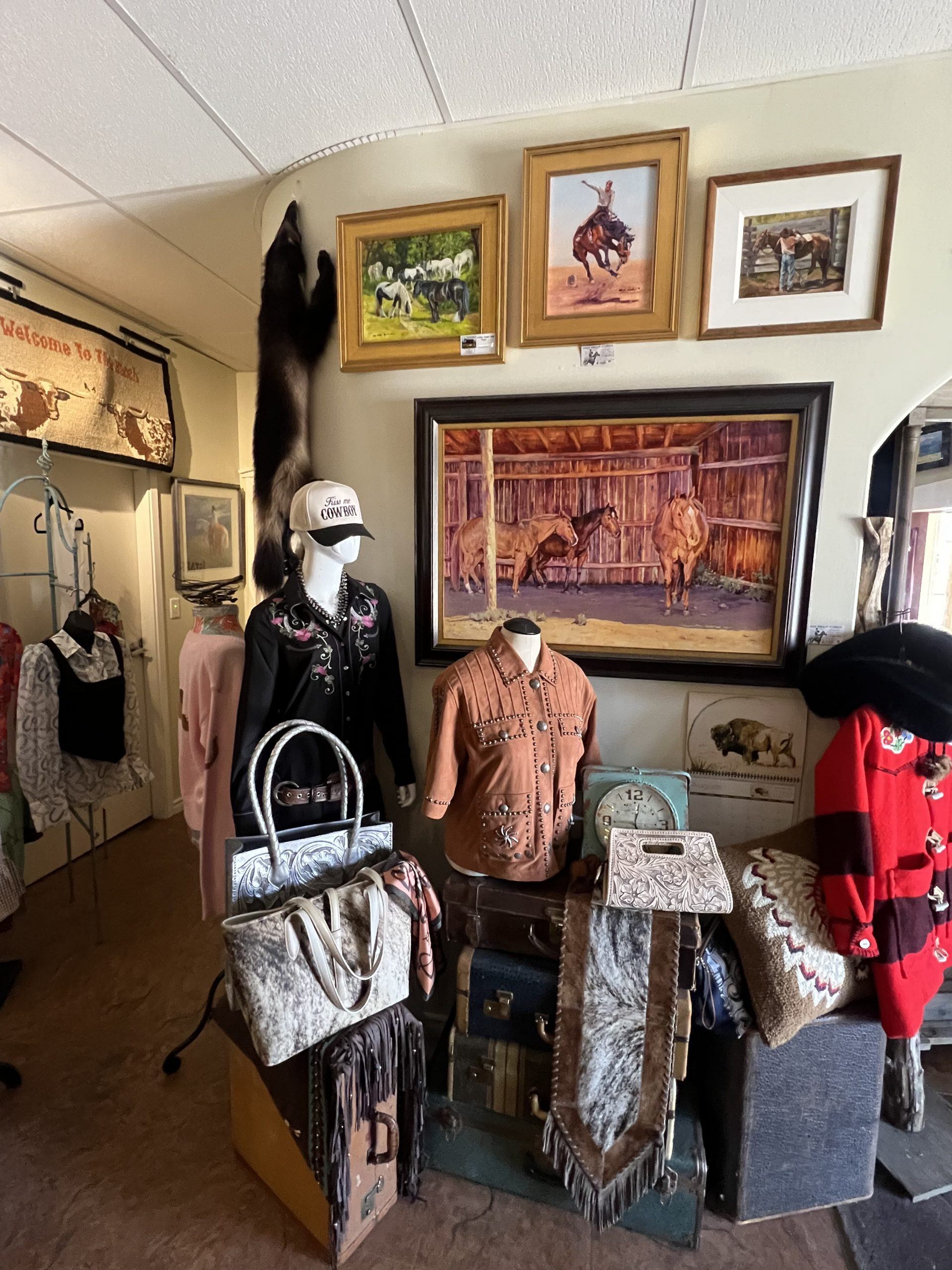 Vintage western clothing display with mannequins, leather bags, fur, and horse paintings on wall.