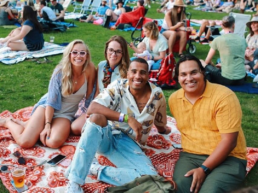 Group of friends sitting on picnic blanket at outdoor event with crowd behind.