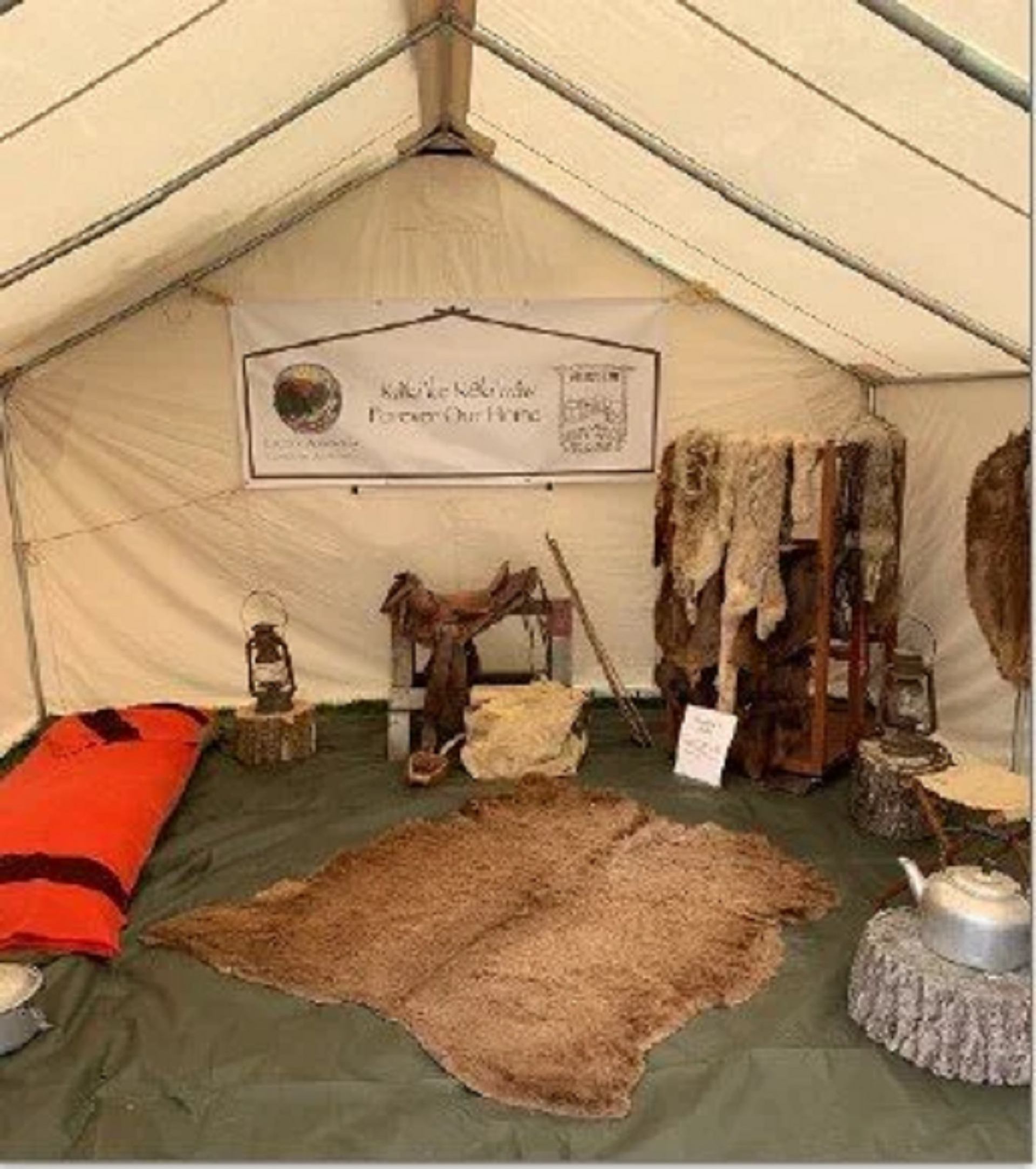 A canvas tent interior with a banner reading "Kallike NèK'ende Forever Our Home," hanging fur pelts, a saddle, and a large hide rug on the floor.